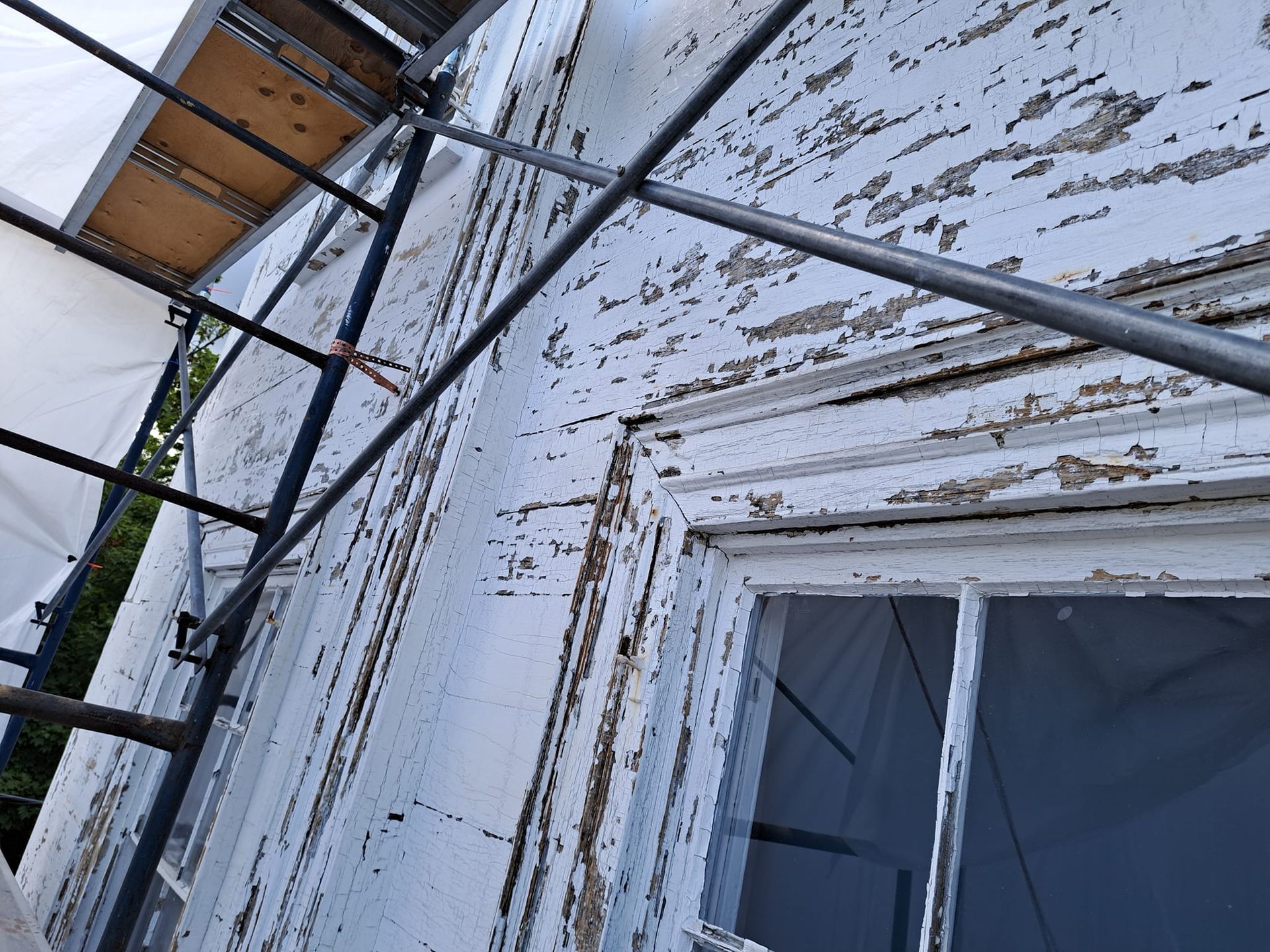 Scaffolding alongside a white house with peeling paint, by a window.