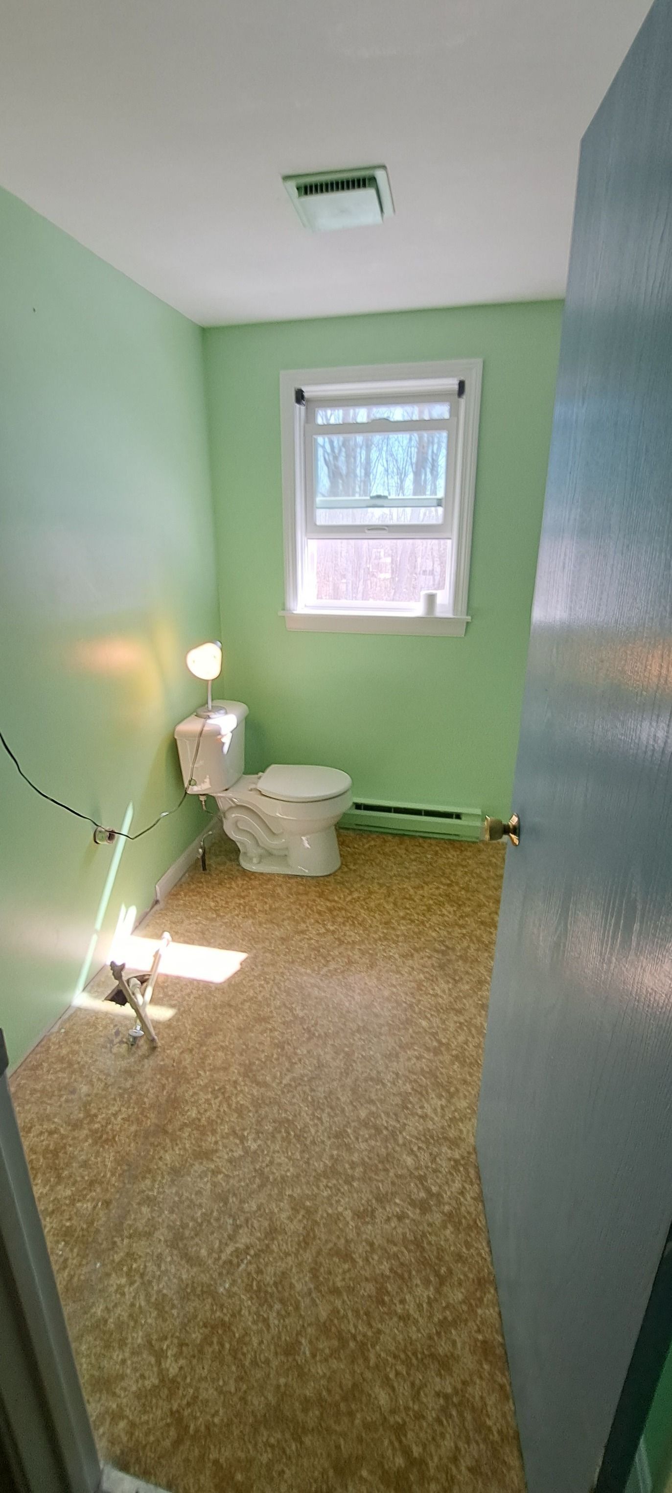 A small, pale green bathroom with a toilet, window, and speckled flooring. A small lamp sits atop the toilet.