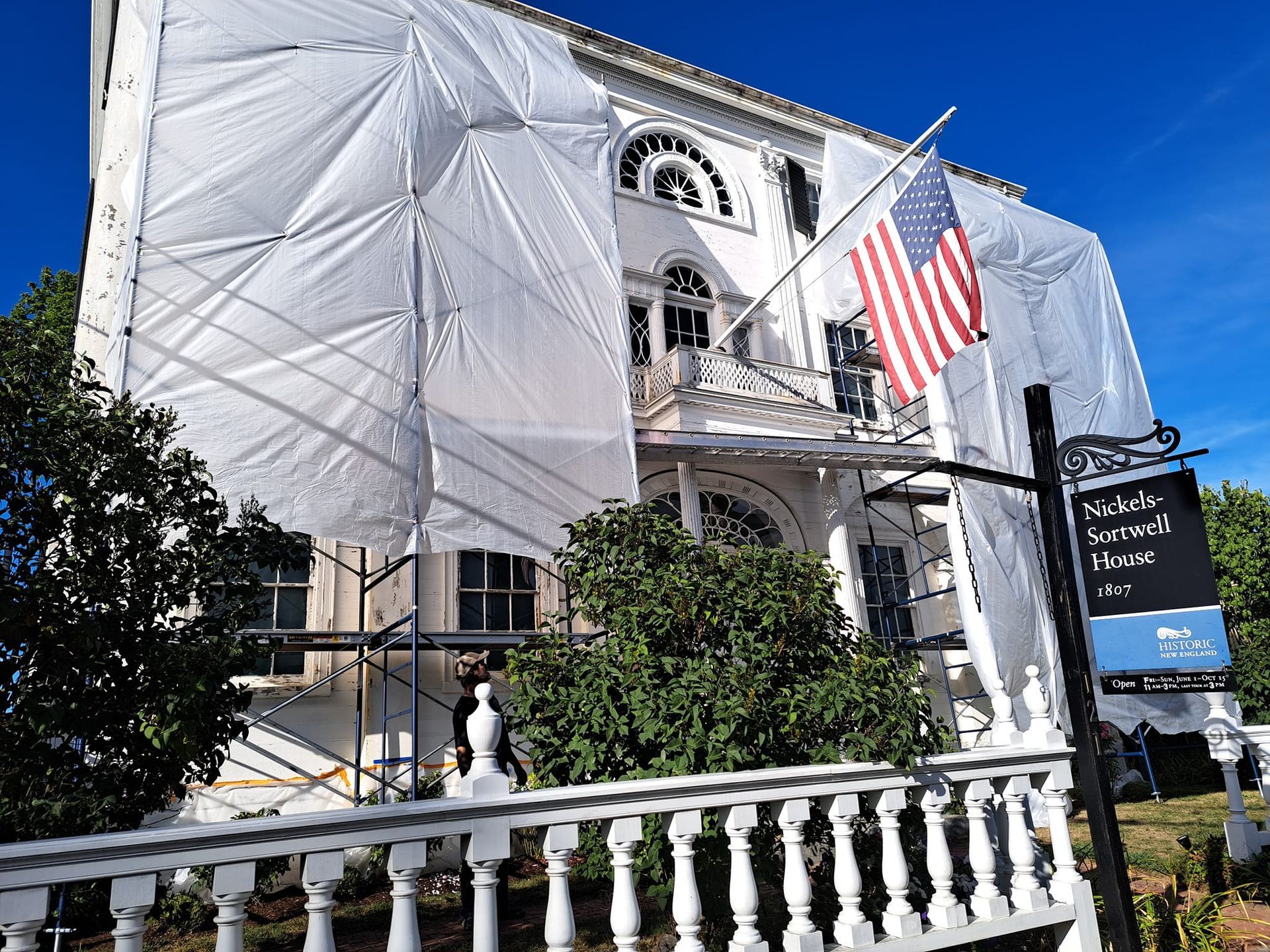 Historic house under renovation, draped in white protective sheeting; American flag flies.