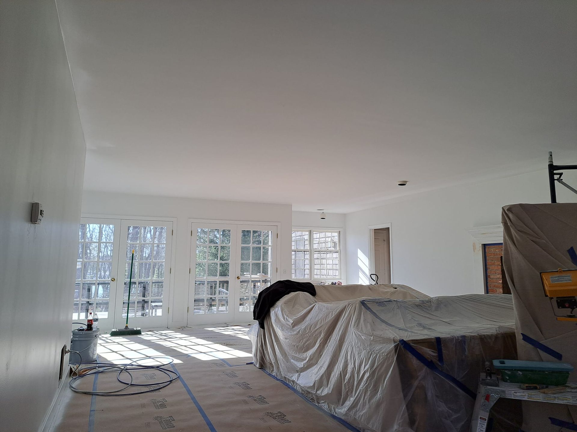 Interior view of a room being painted, featuring French doors, covered furniture, and a partly painted ceiling.