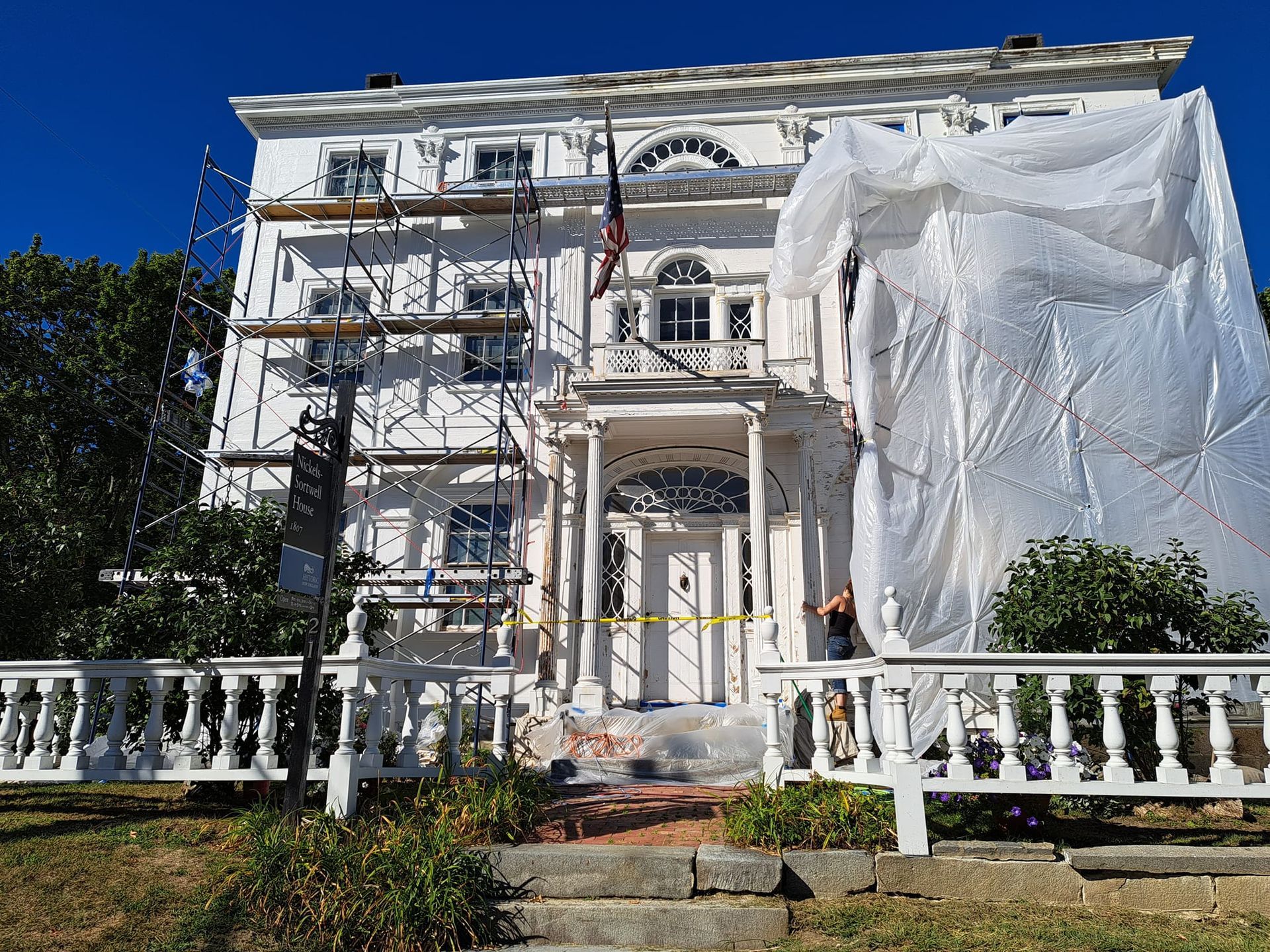 White building with scaffolding and tarp for renovations. American flag flies. Yellow caution tape.