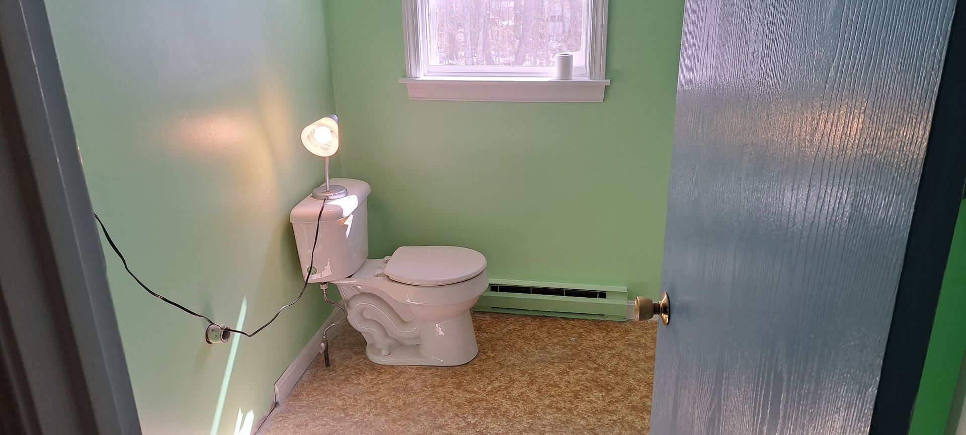 A small, light green bathroom with a toilet, window, and speckled flooring. A door is partially open.