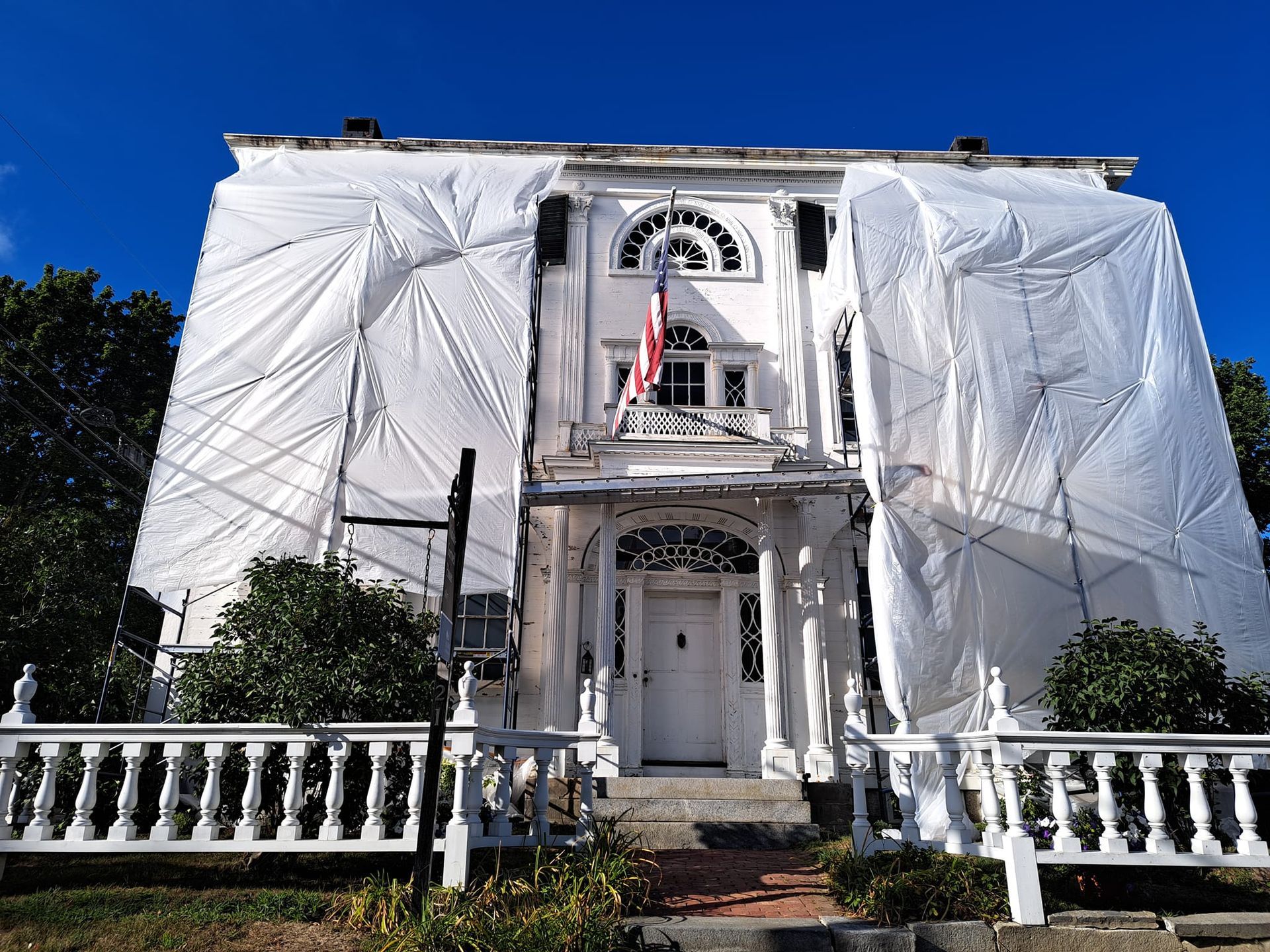 White historic house, scaffolding and covering, American flag, blue sky, white fence.