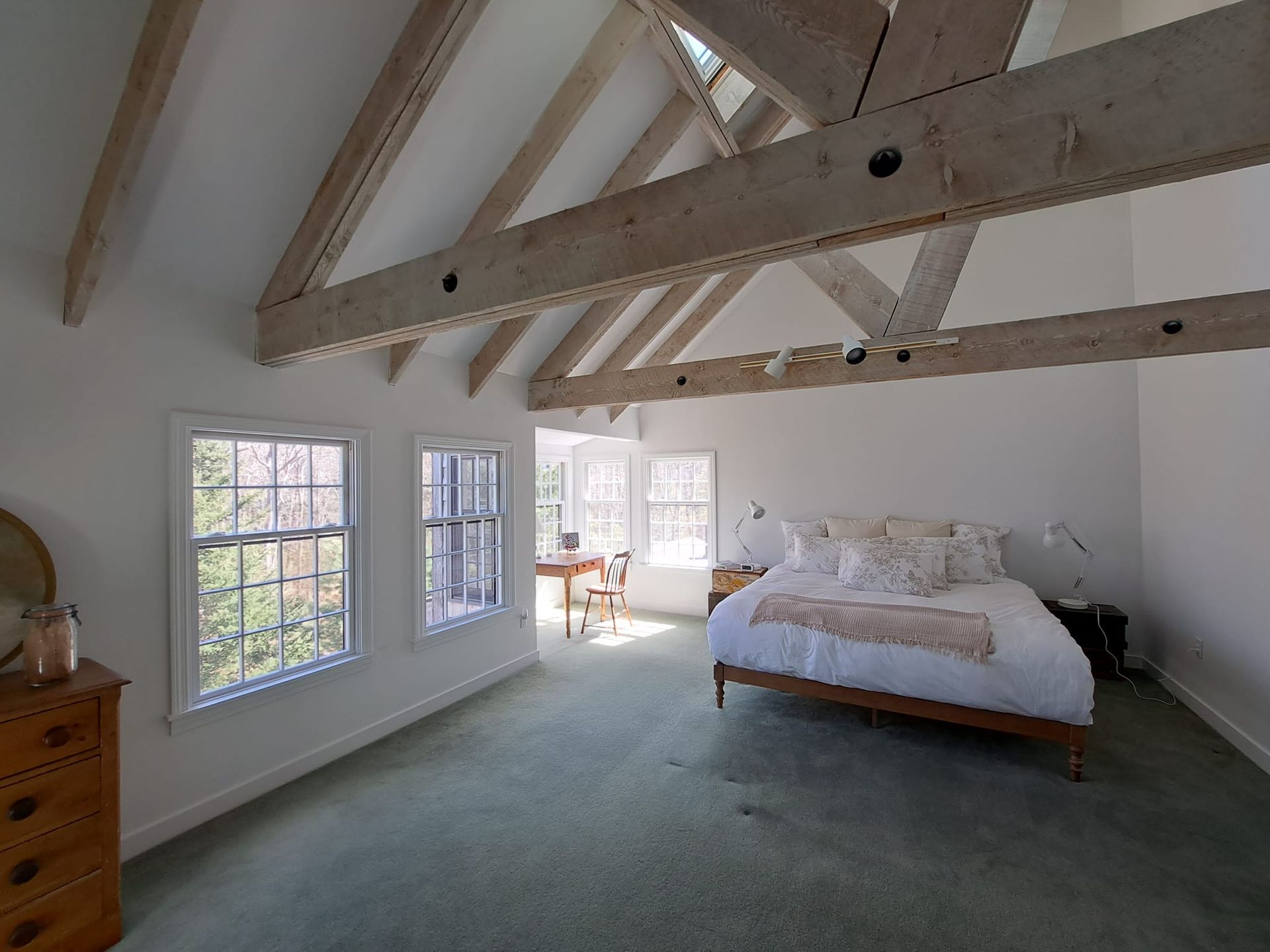 Bedroom with white walls, green carpet, wood beams, bed, windows, and dresser.