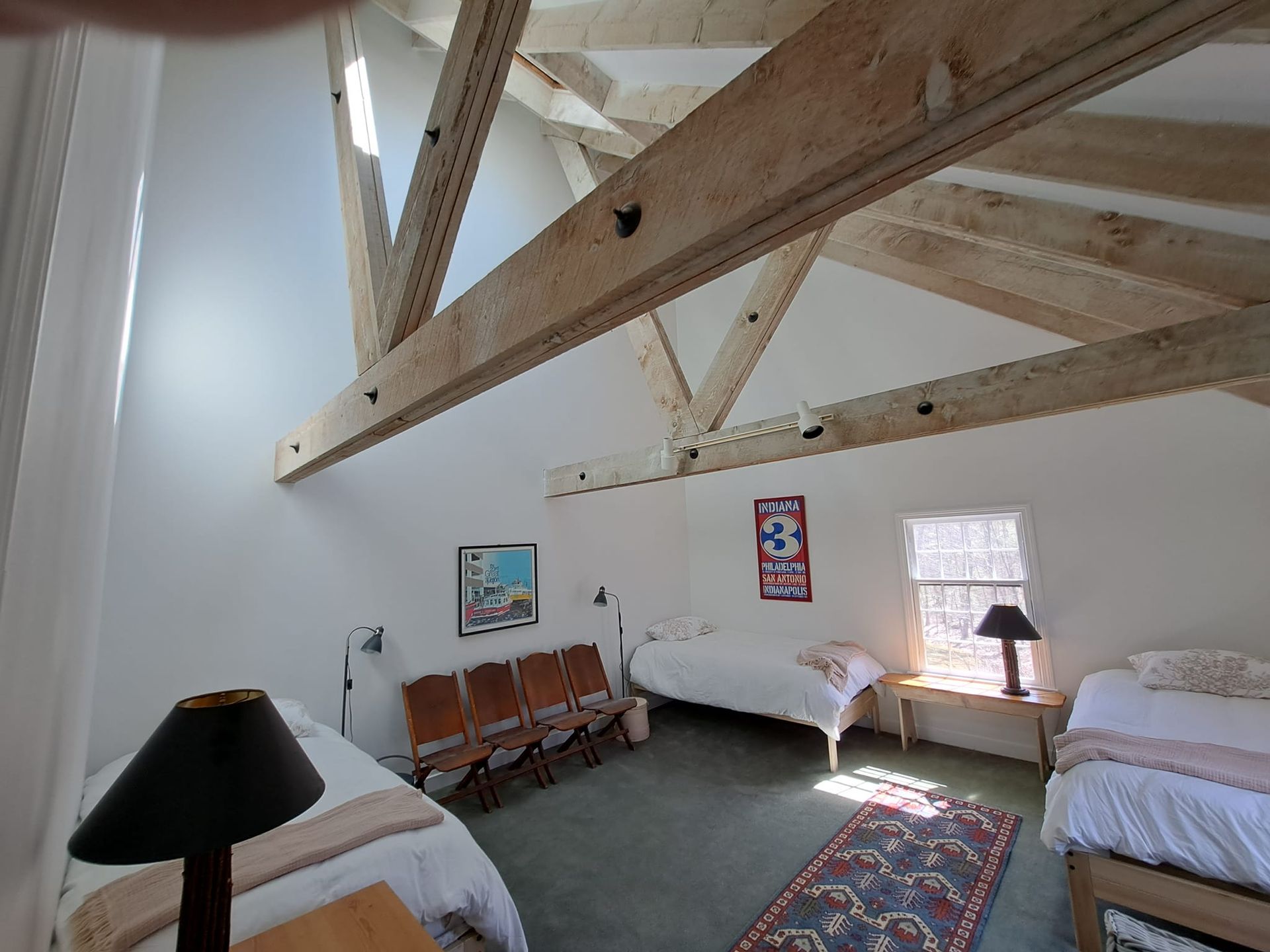 Bedroom with exposed wooden beams, three beds, and a rug. White walls, natural light.