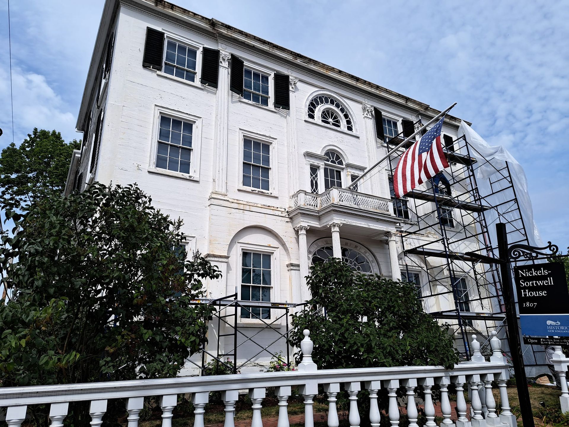 White, three-story building with black shutters and an American flag. Scaffolding visible, likely under renovation.