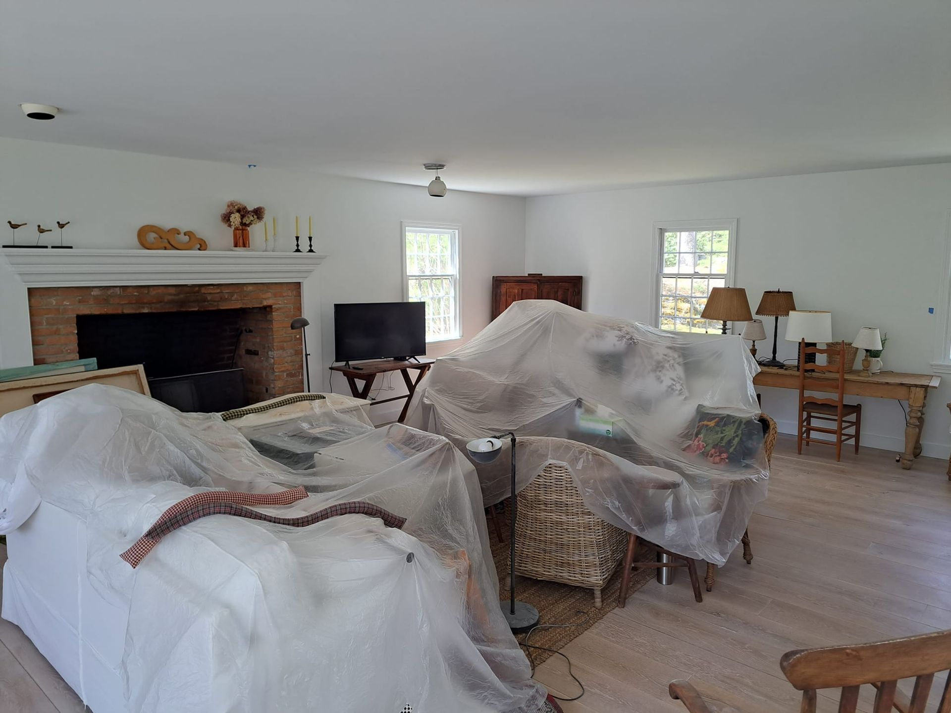 Living room with furniture covered in plastic for protection. Fireplace, windows, and lamps are visible.