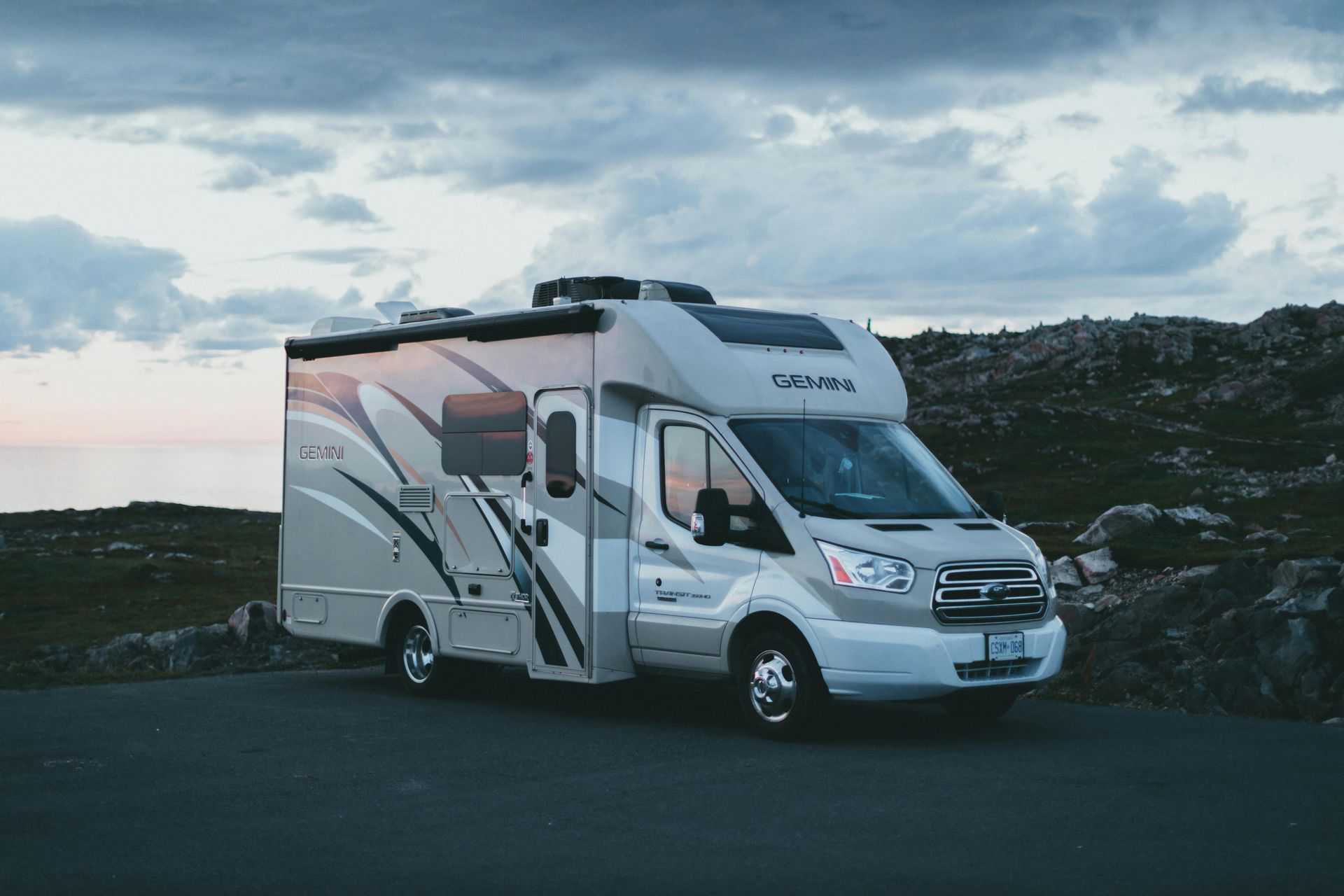 RV parked on asphalt road, rocky landscape, cloudy sky.