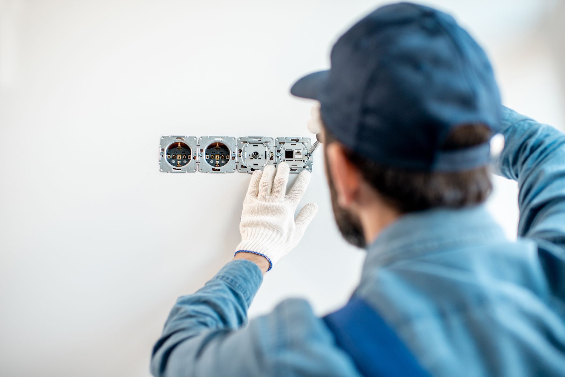 Electrician installing an electrical outlet on a white wall, wearing gloves and a cap.
