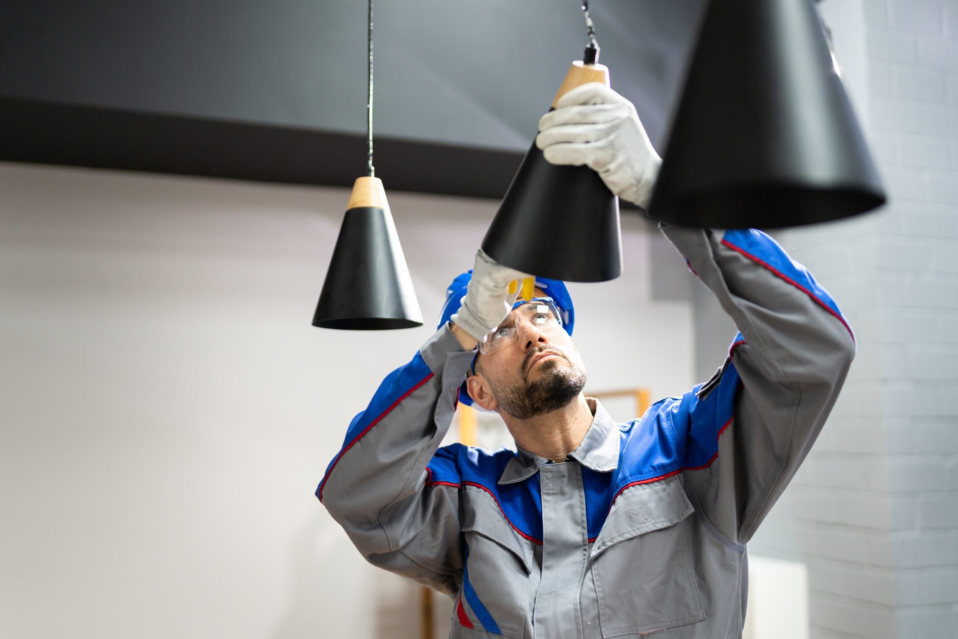 Electrician in a blue uniform installing a black pendant light fixture indoors.