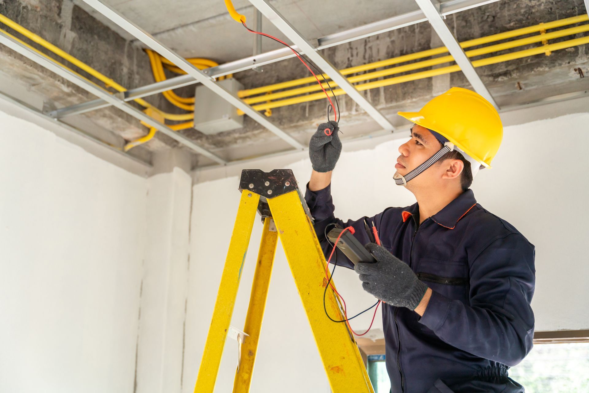 Electrician on a yellow ladder, testing wires in a building's ceiling, wearing a yellow hardhat and gloves.