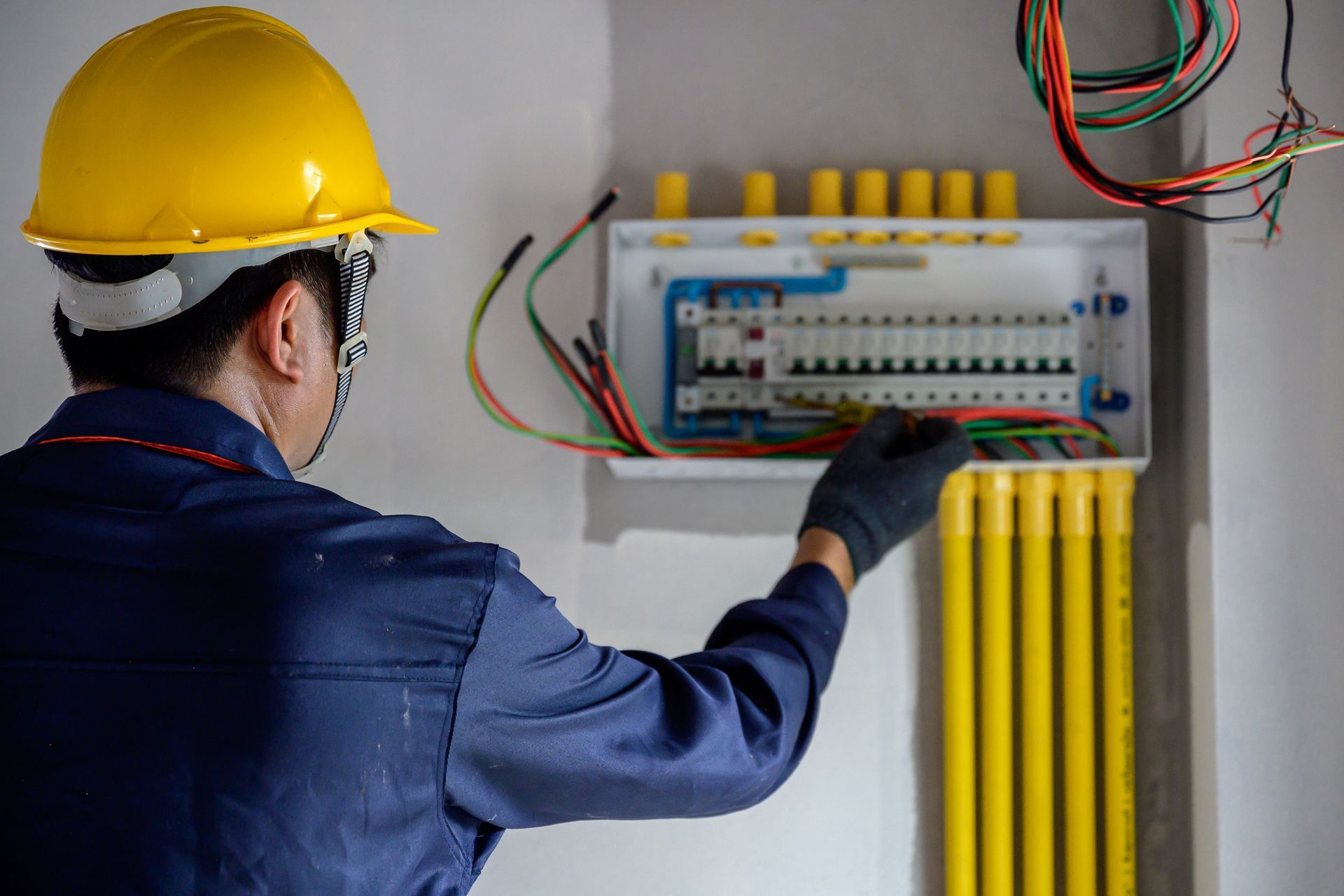Electrician working on electrical panel, wearing yellow hard hat and blue jumpsuit.