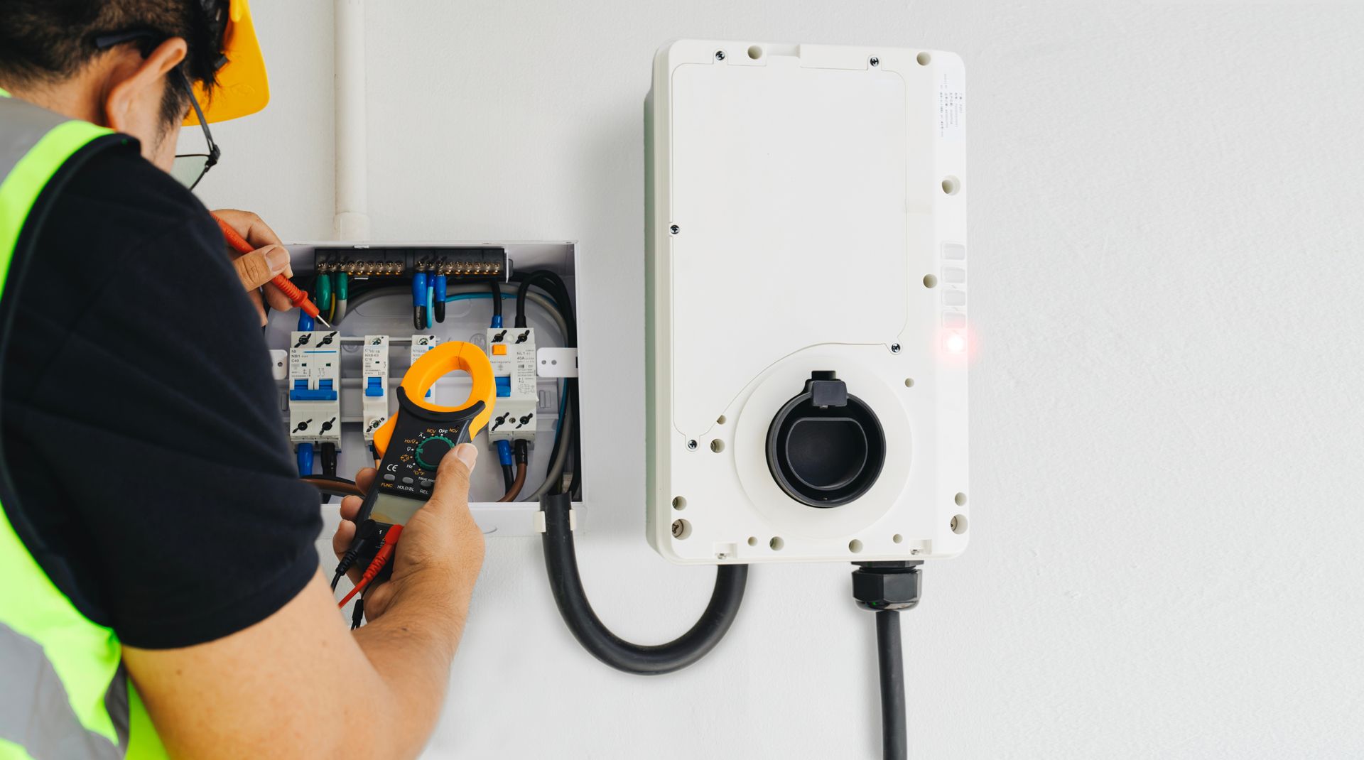 Electrician using a clamp meter to work on a white electrical box attached to a wall.