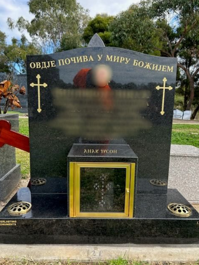 Black granite tombstone with gold crosses, inscription, and a small memorial glass box.