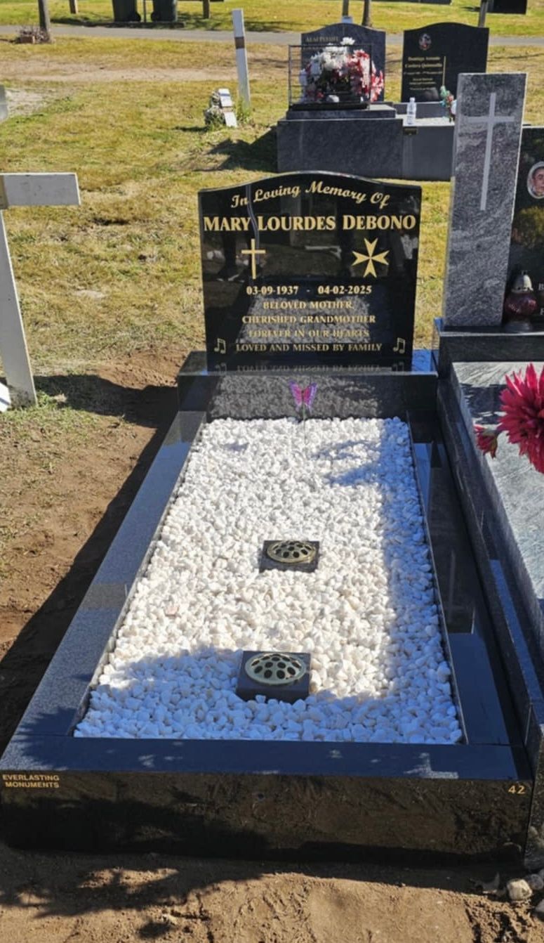 Black granite headstone in a cemetery with white gravel and a cross.