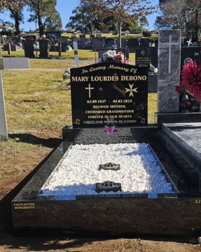 Headstone for Mary Lourdes Debono in a cemetery, black with gold lettering, surrounded by white stones.
