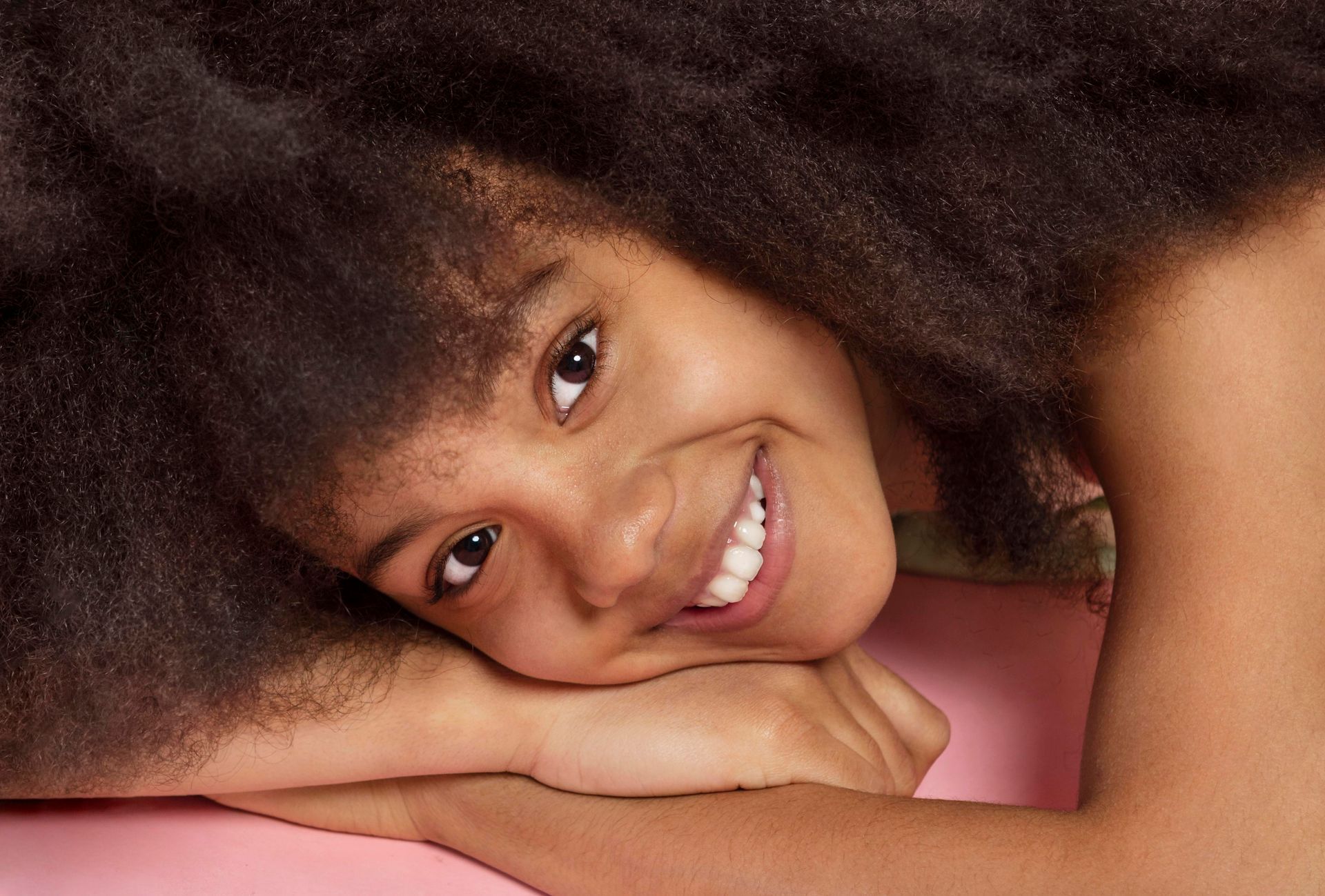 A woman with an afro is smiling while laying on her stomach on a pink surface.