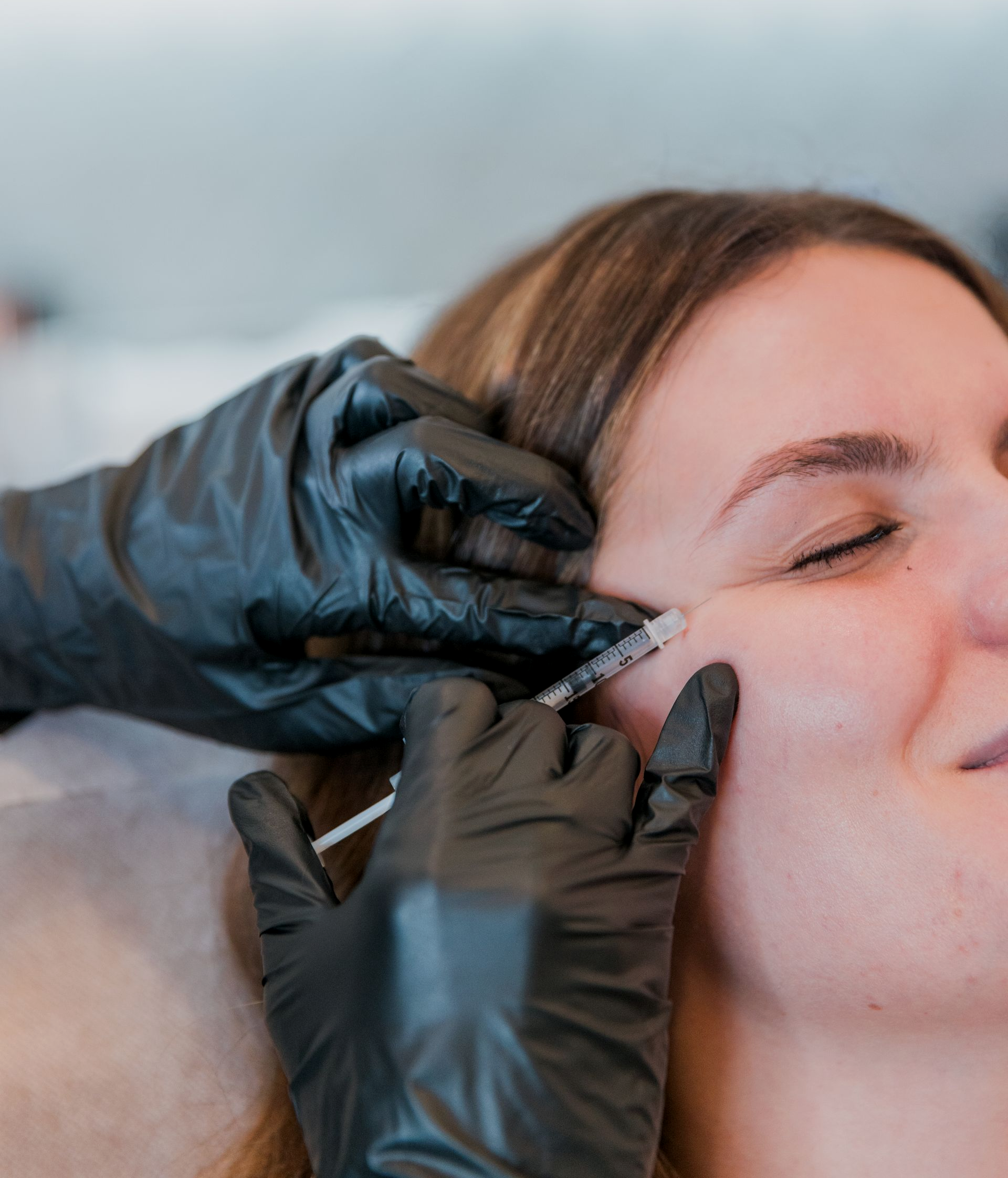 Person receiving a facial injection near the eye, gloved hands hold syringe.