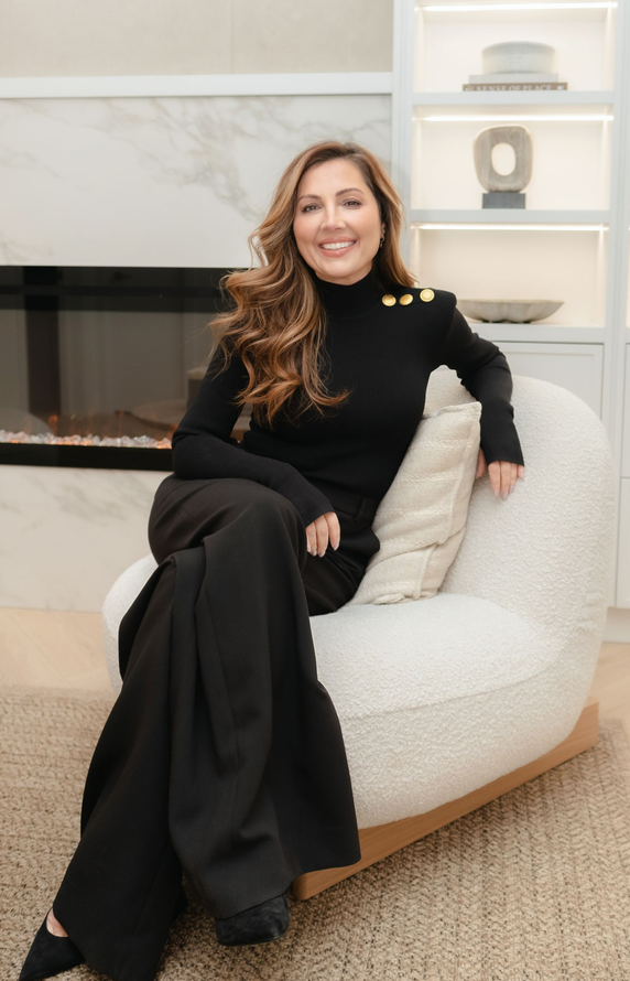 A woman is sitting on a stool wearing a white sweater and pants.