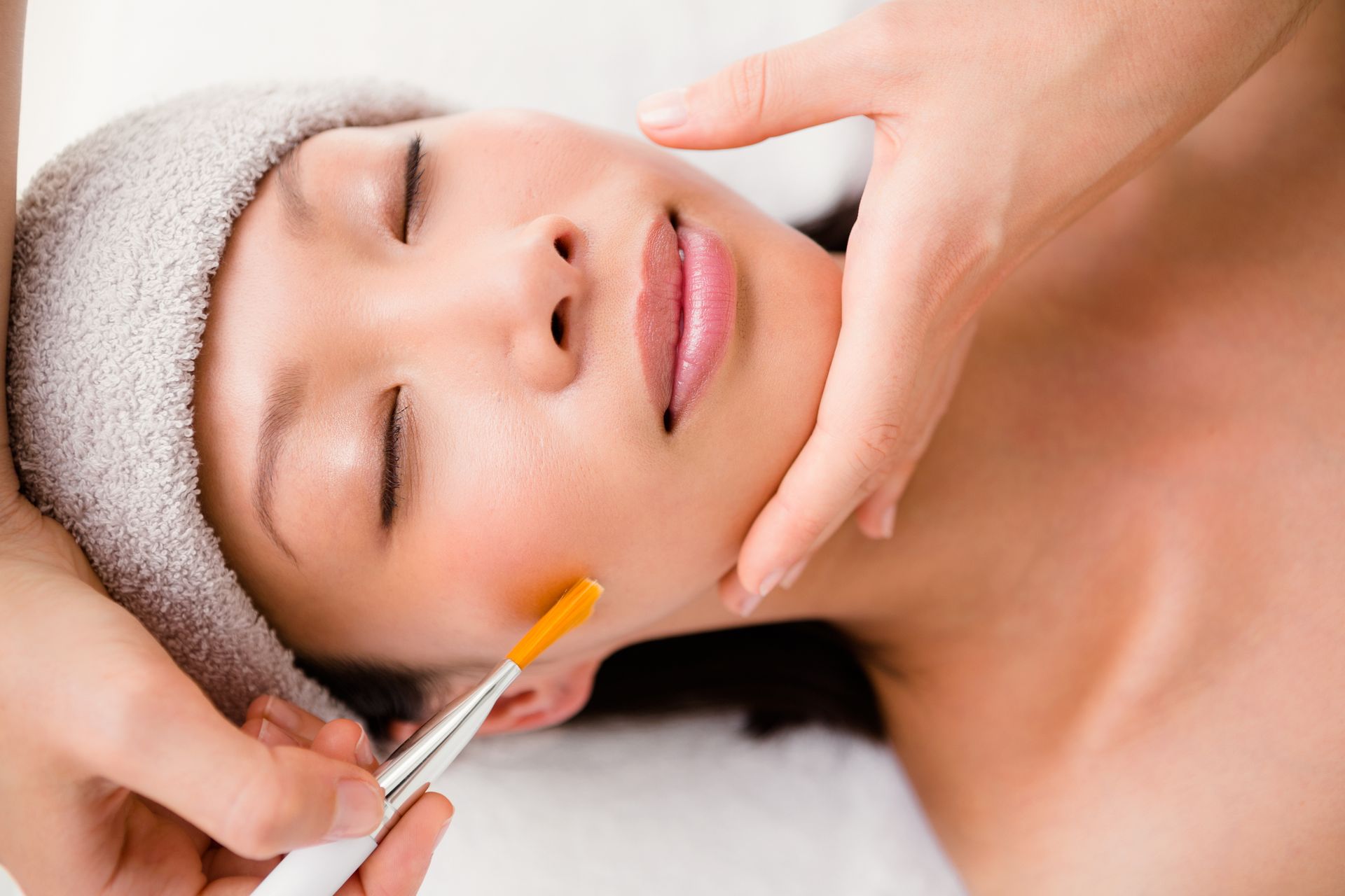 Woman receiving a facial treatment; brush on cheek, eyes closed, spa setting.