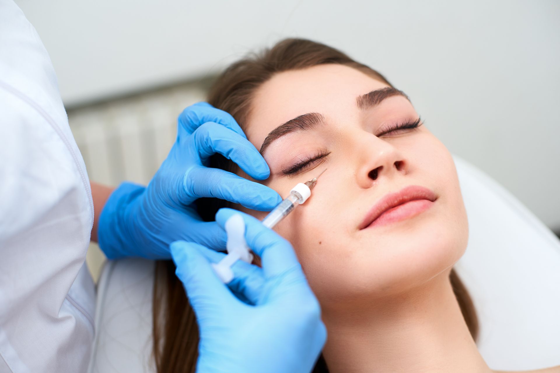 A person receiving a cosmetic injection near the eye, blue gloves, neutral setting.