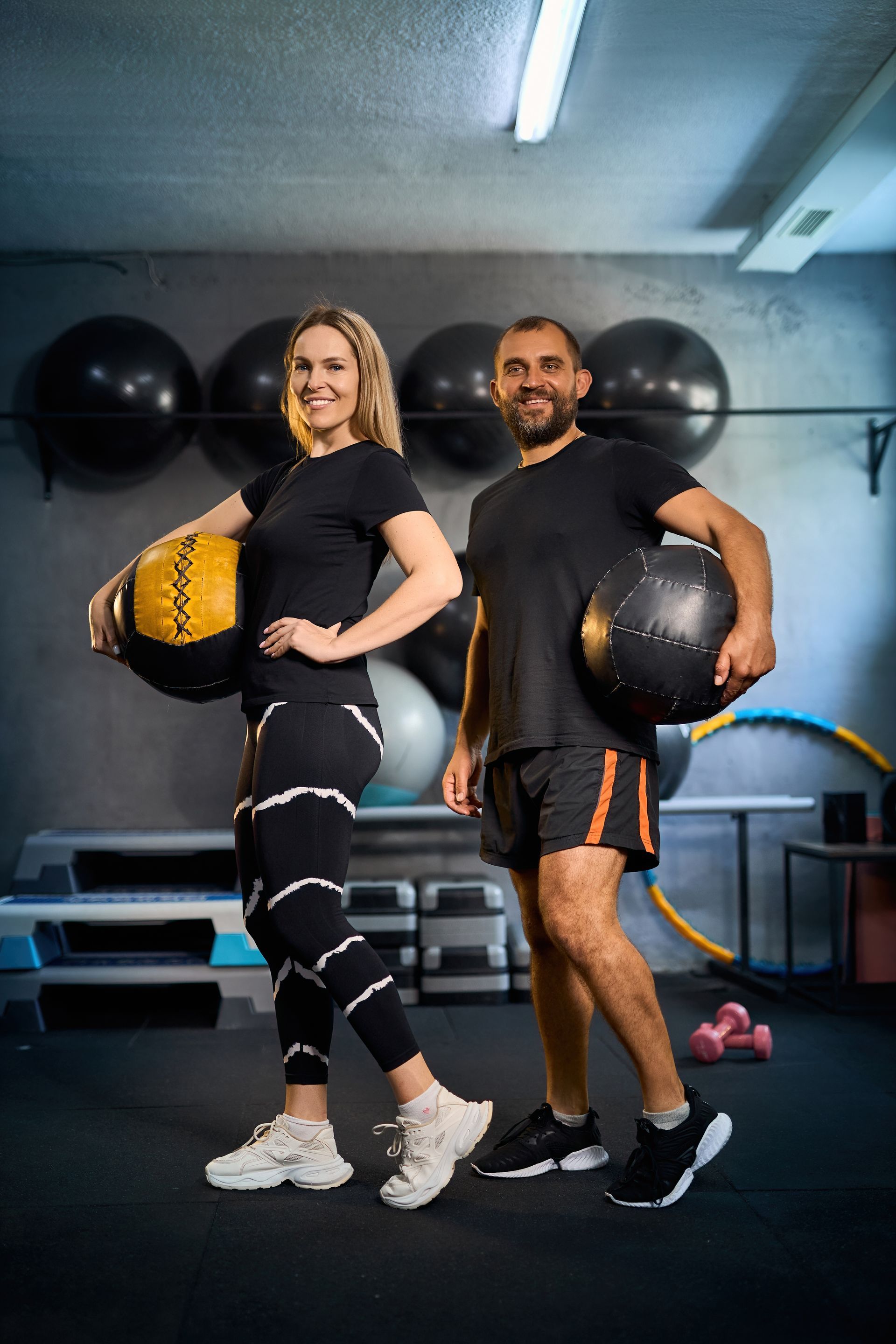 A man and a woman are standing next to each other in a gym holding medicine balls.