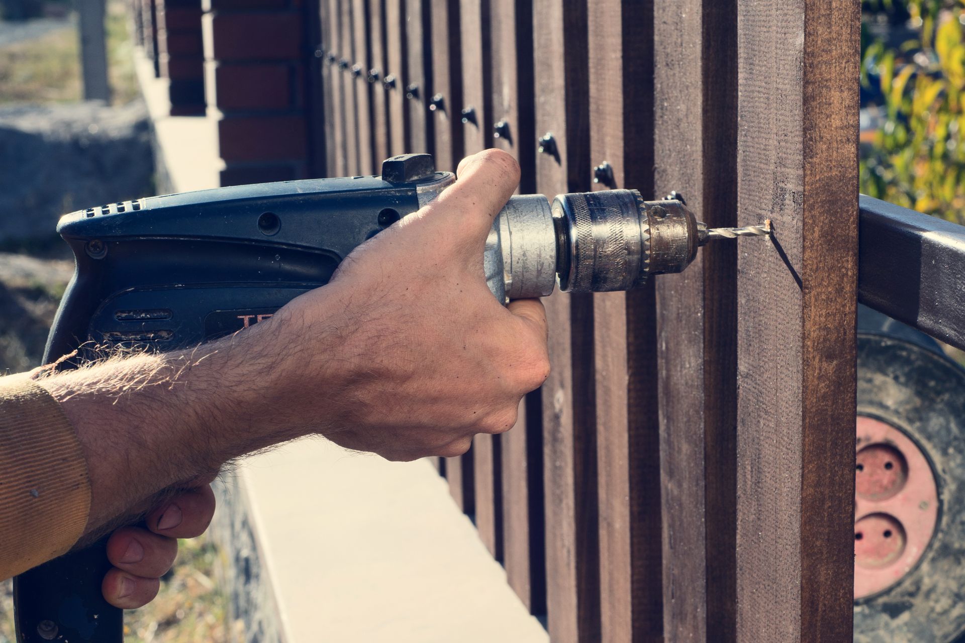 Close-up of a hand using a power drill to fasten a screw into a wooden fence.
