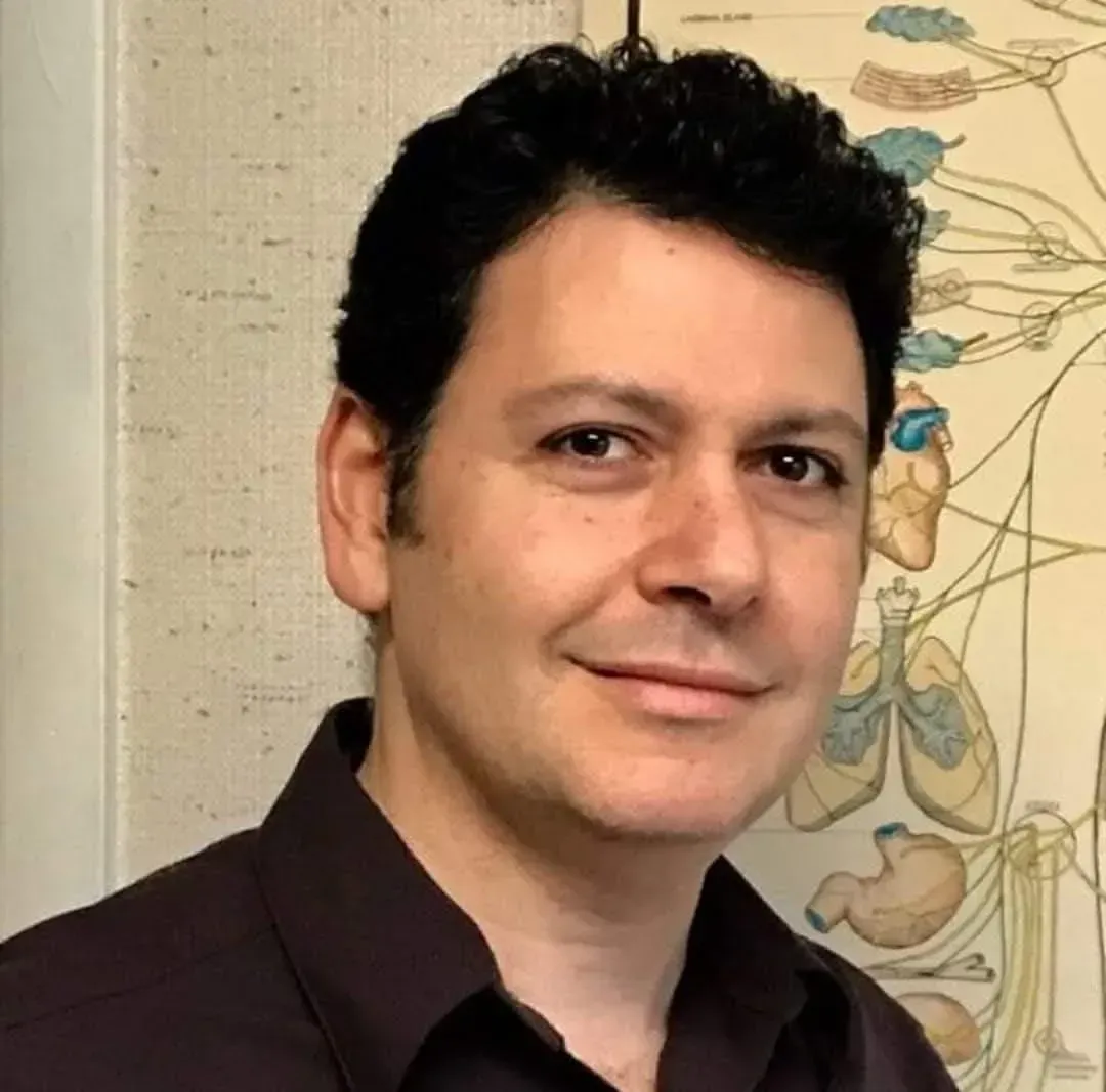Man with dark hair and a slight smile, standing near a medical chart. He wears a dark button-down shirt.