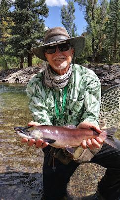 Man holding a fish by a river, wearing a hat and sunglasses, smiling.