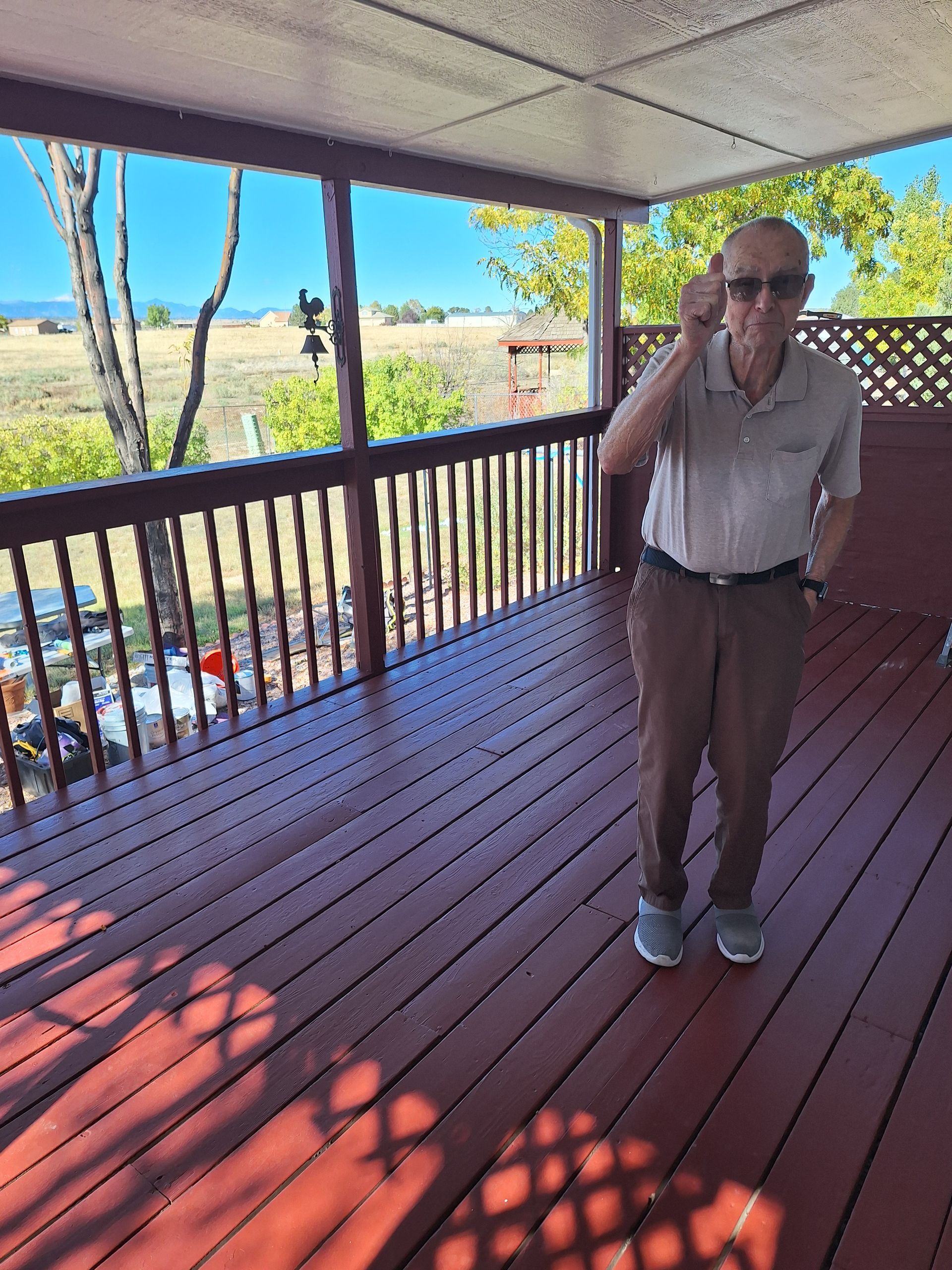 Man on a red wooden deck, holding up sunglasses. Overlooks a field on a sunny day.