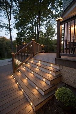 Wooden deck with stairs lit by small lights, leading to a house, against a backdrop of trees.