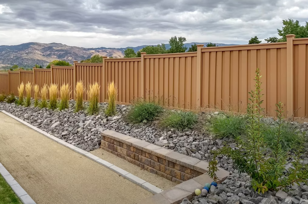 Wooden fence along a rocky bed with decorative plants and a tan path, mountains in the background under a cloudy sky.
