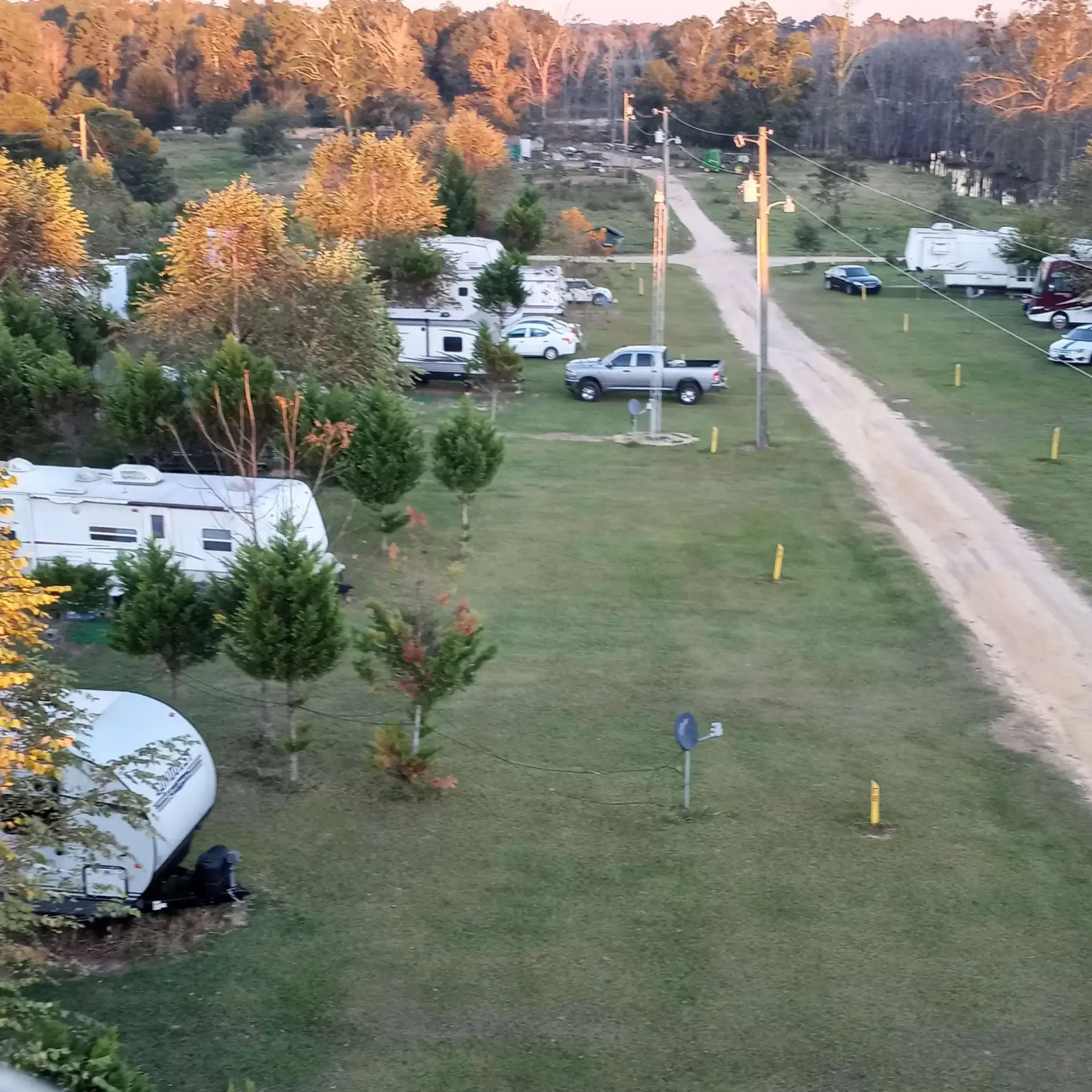 RV campground with trailers parked on grassy plots, dirt road. Trees in background, sunset.