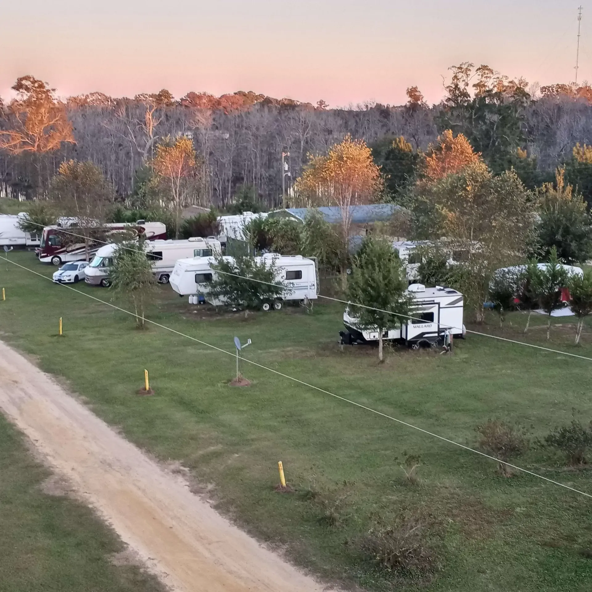 RV campground at dusk. Several recreational vehicles parked on green grass, dirt road in foreground, trees in background.