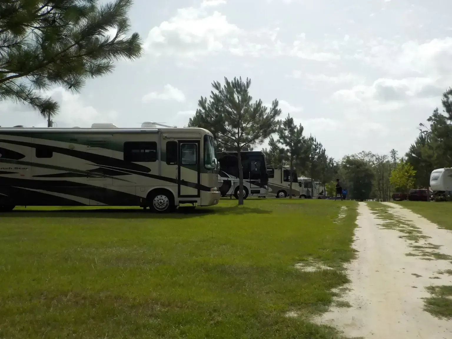 RVs parked in a grassy campground, a dirt path leads through the sites under a cloudy sky.