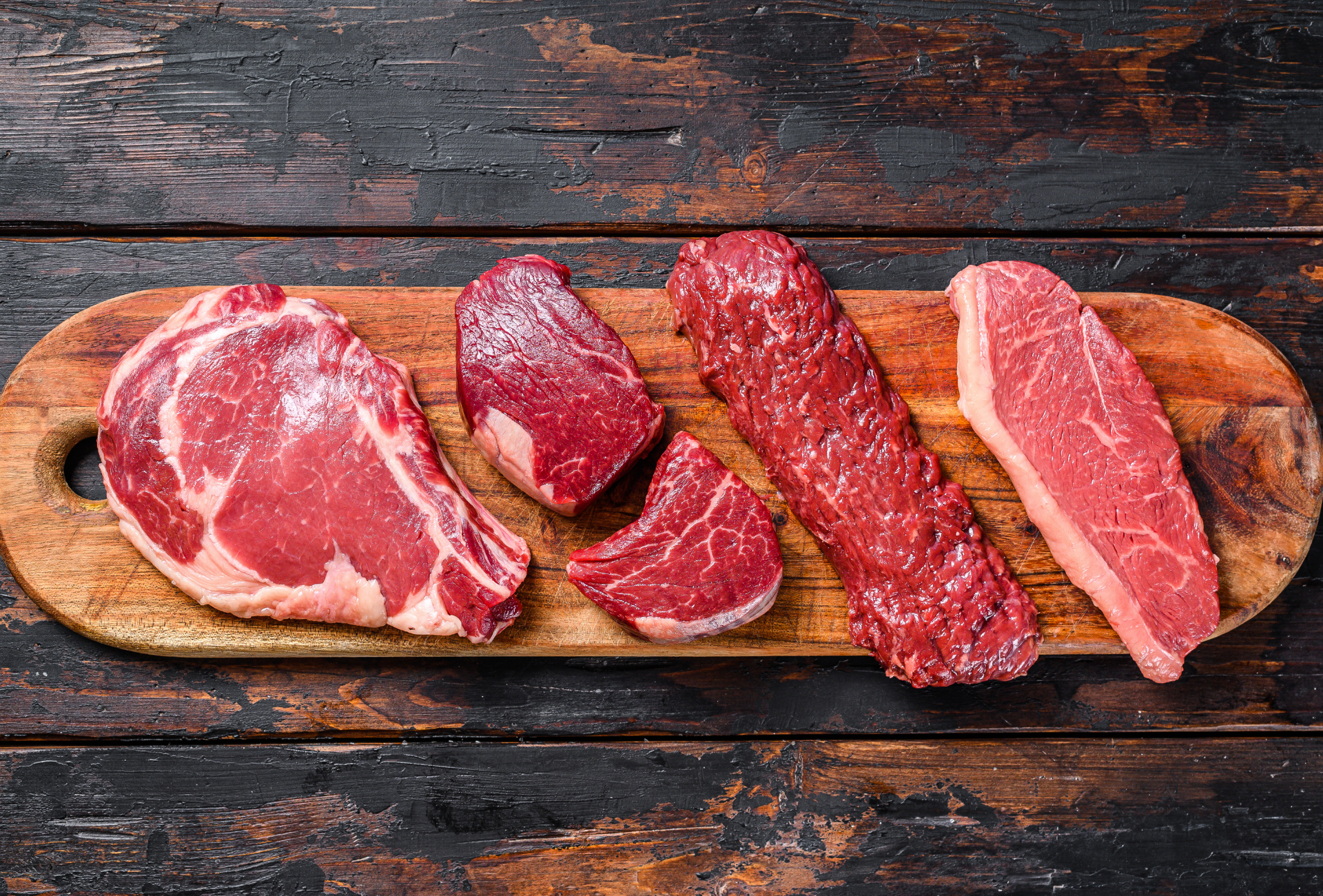Raw cuts of beef on a wooden cutting board, set on a dark wooden table.