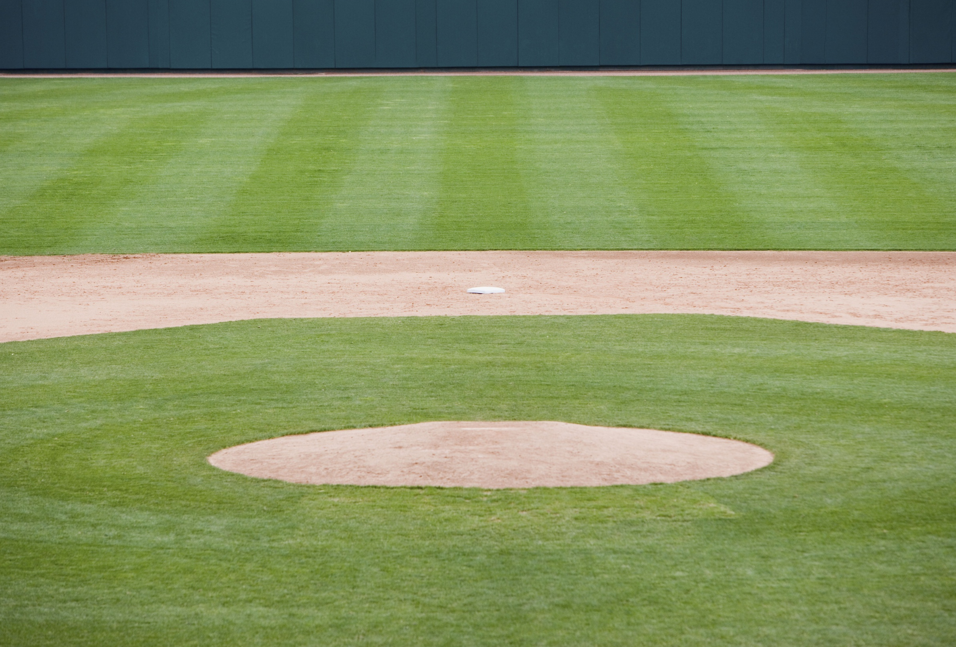 Baseball field view, green grass, dirt mound and base, green outfield wall.