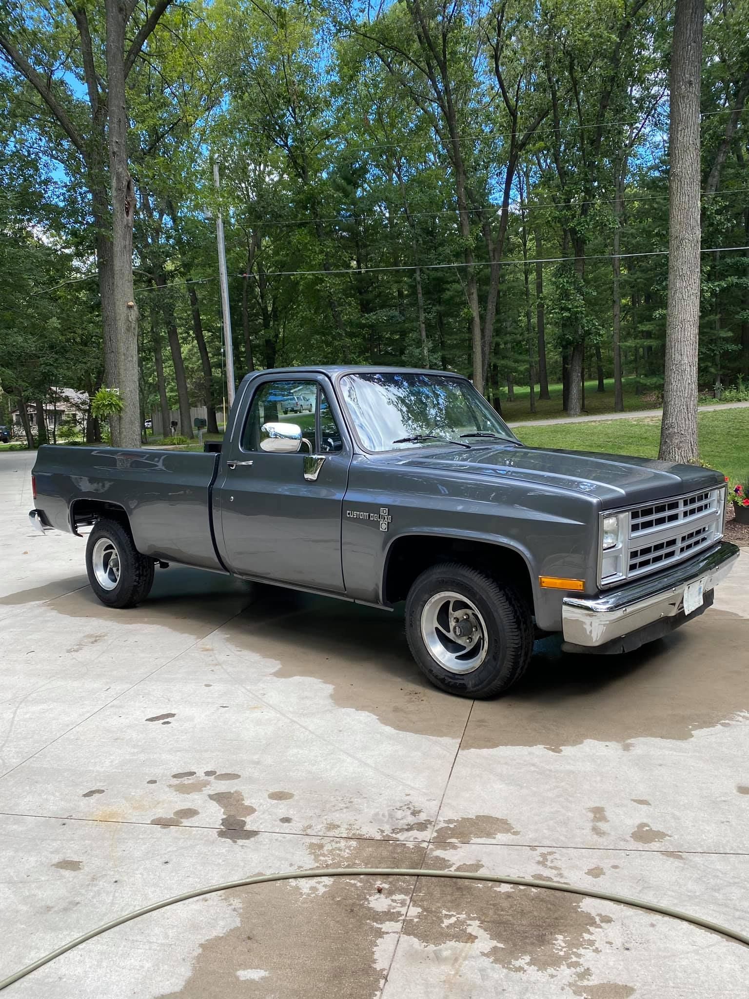 Gray Chevrolet pickup truck parked on a concrete driveway, surrounded by trees and a grassy yard.