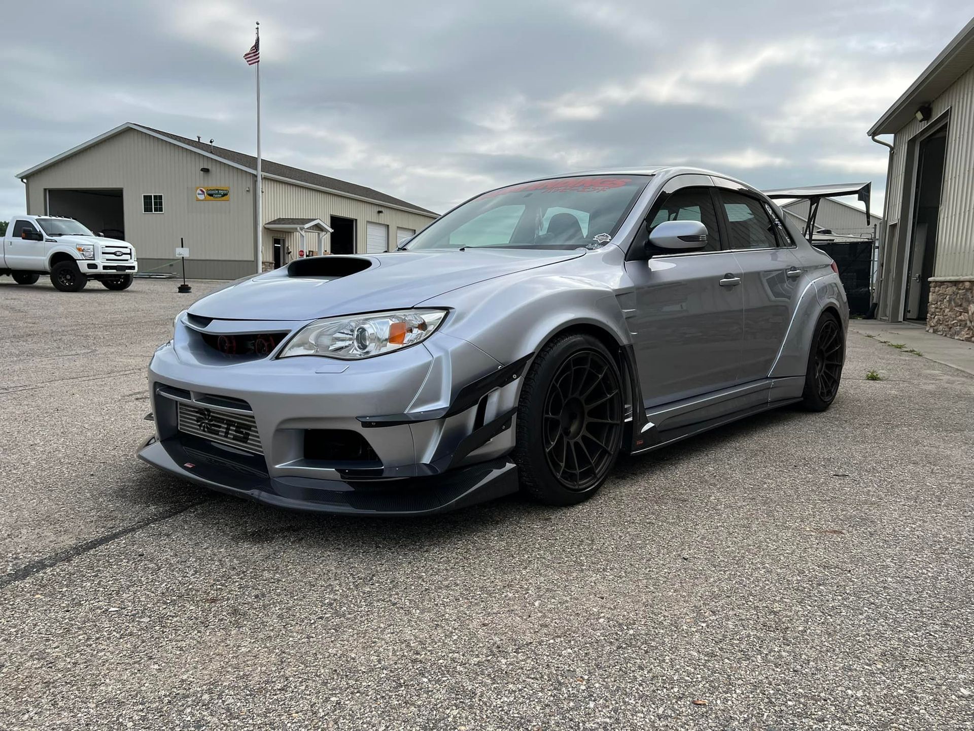 Silver Subaru with wide fenders and black wheels parked in front of a building on a cloudy day.