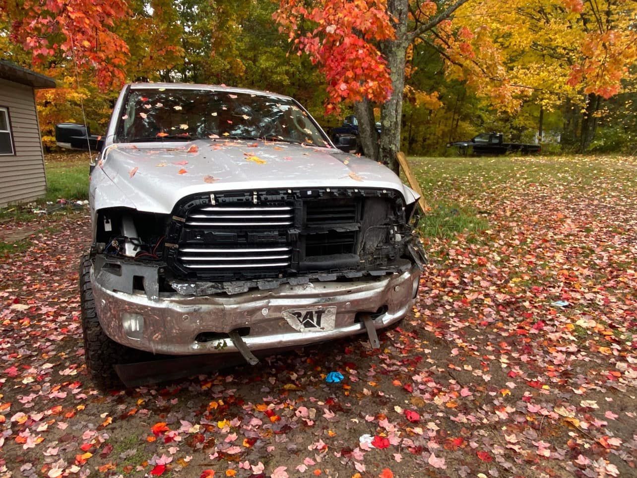 White pickup truck with front-end damage parked on a leaf-covered lawn beneath autumn foliage.