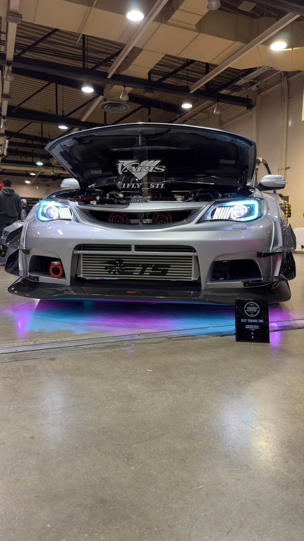 Gray Subaru with hood open, blue and purple underglow, at a car show.