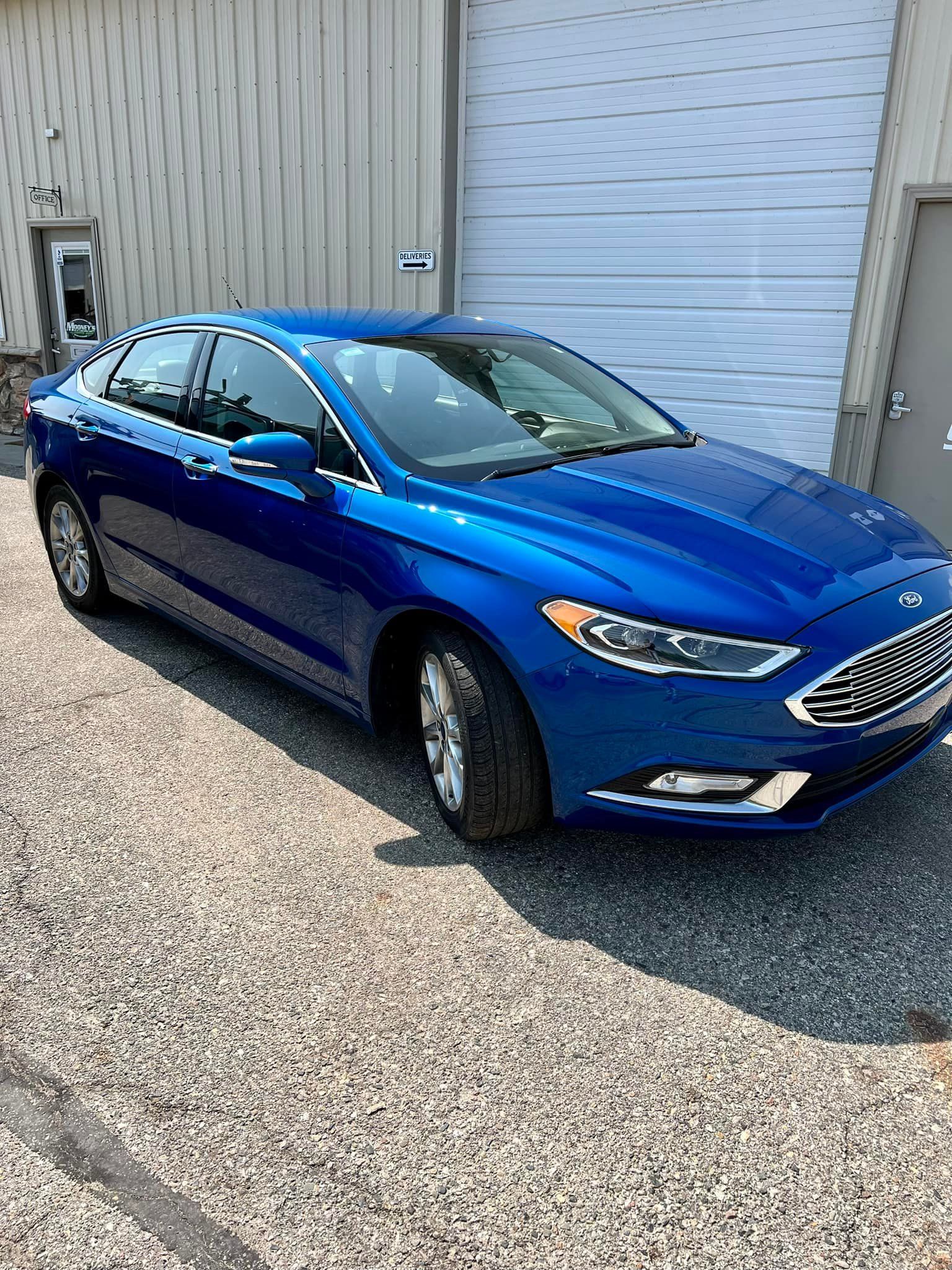 Blue Ford Fusion parked in front of a building with a closed garage door on a gravel surface.