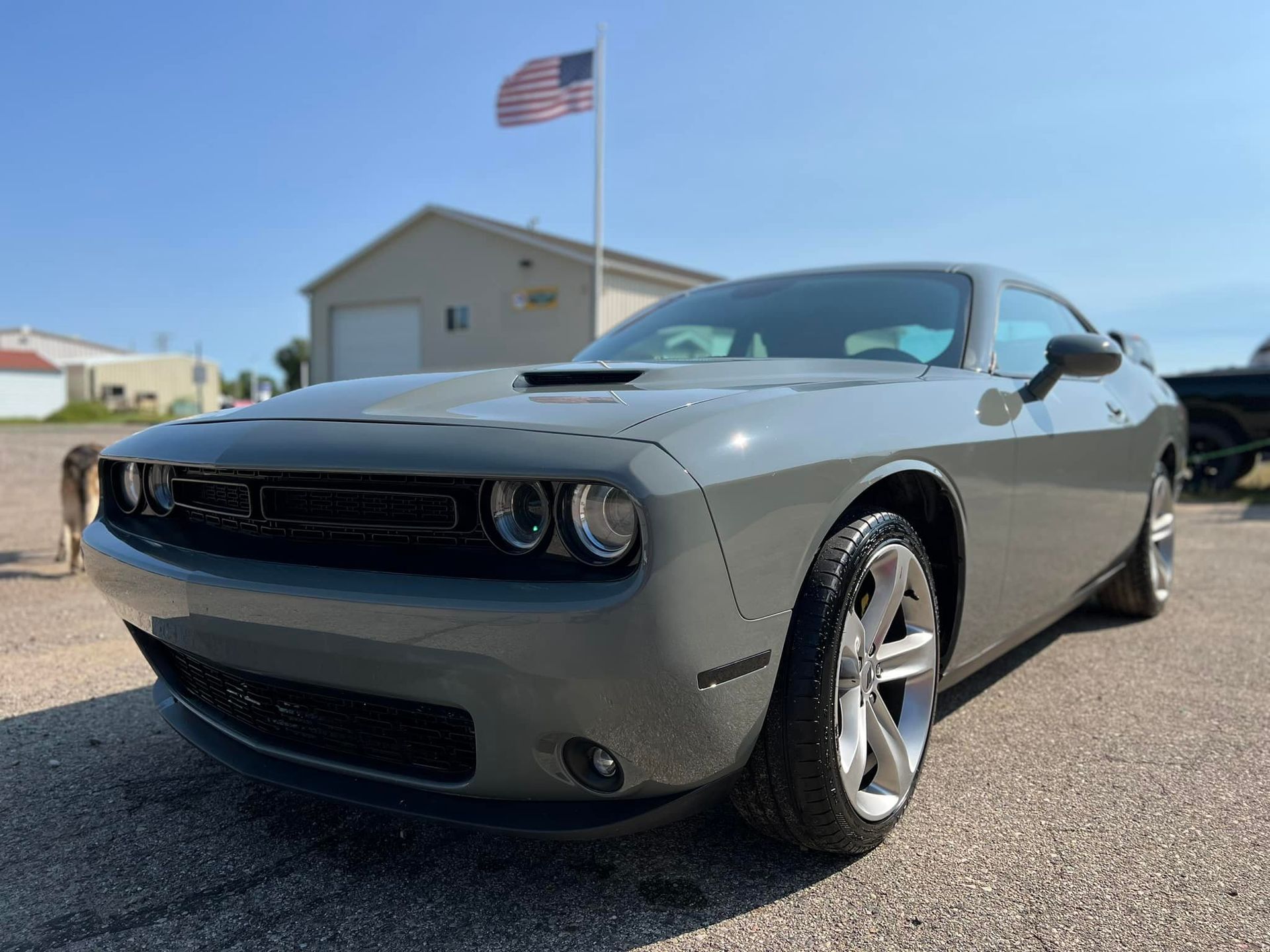 Gray Dodge Challenger car parked in front of a building with an American flag on a sunny day.