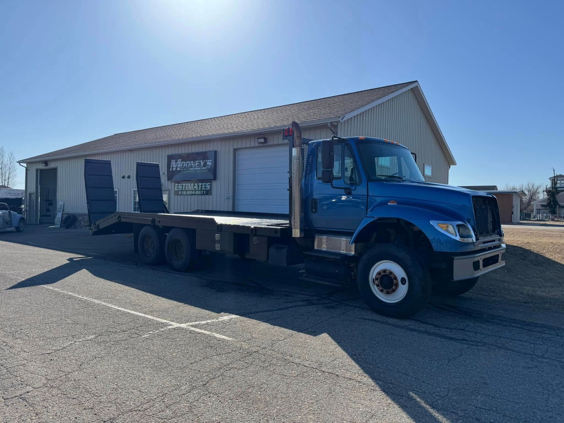 Blue flatbed truck parked outside a building with a white garage door. Sunny day.