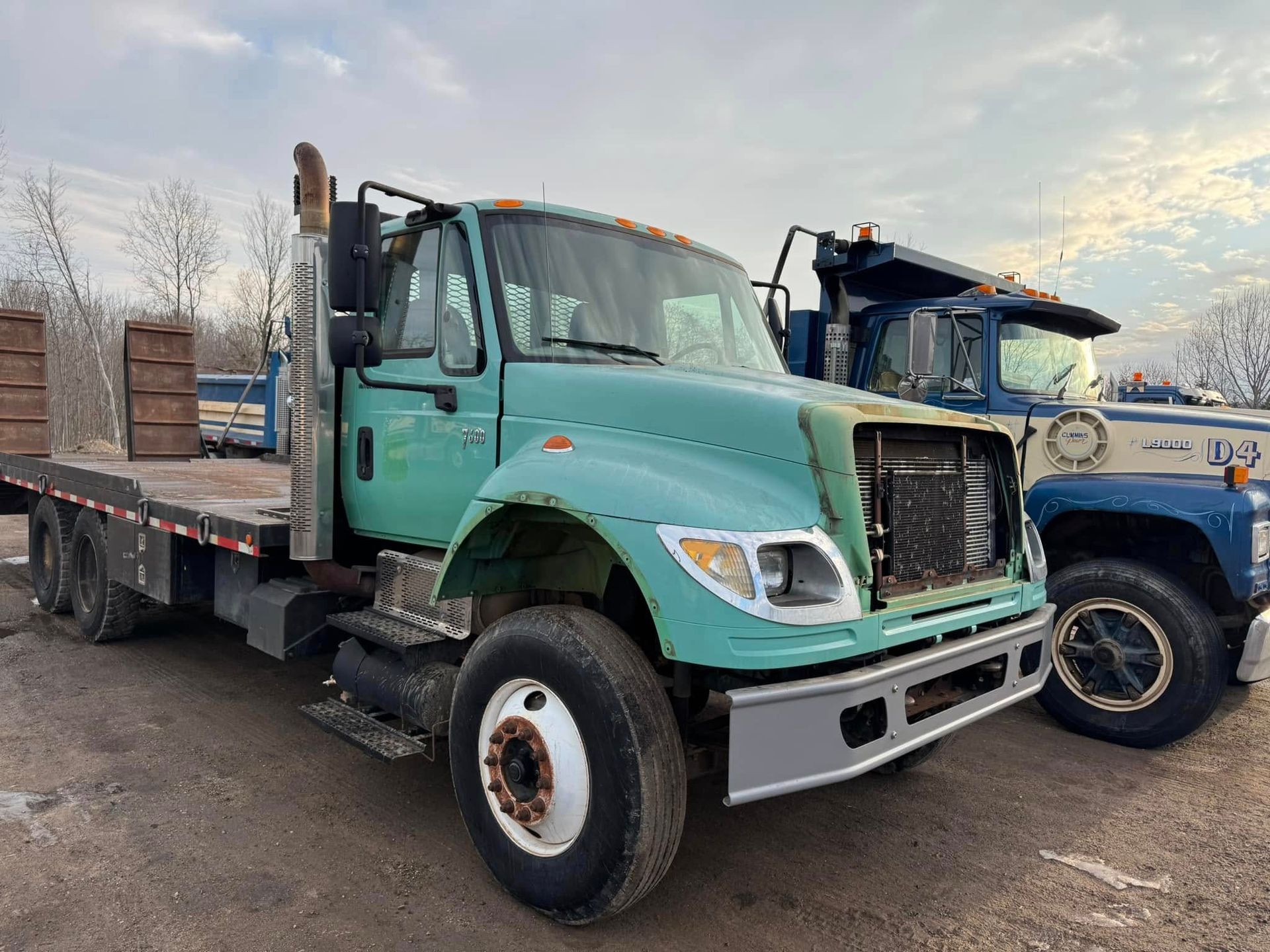 A weathered teal flatbed truck parked next to a blue and white dump truck. Outdoor setting.