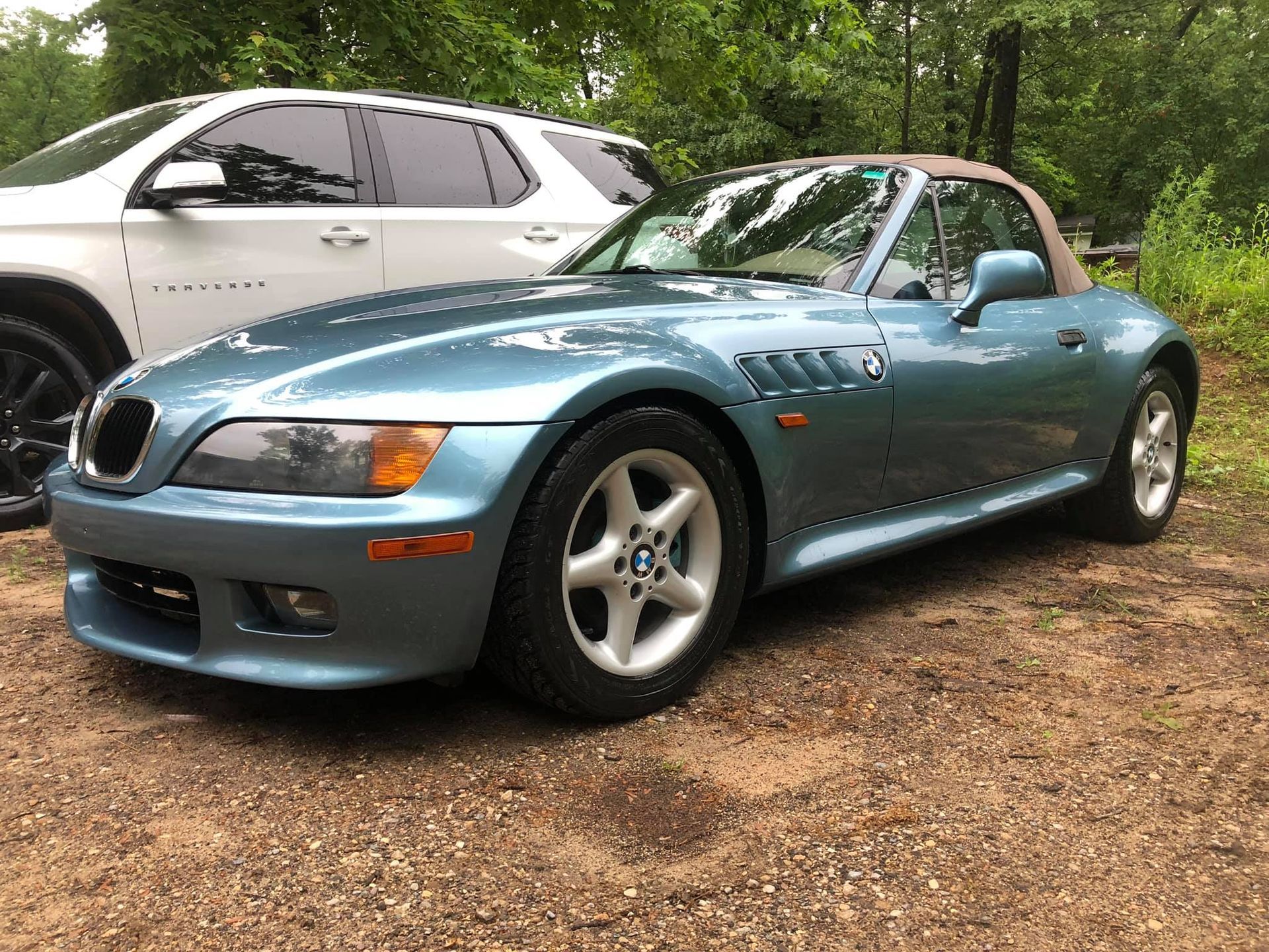 Blue BMW Z3 convertible parked on gravel, with a white SUV in the background.