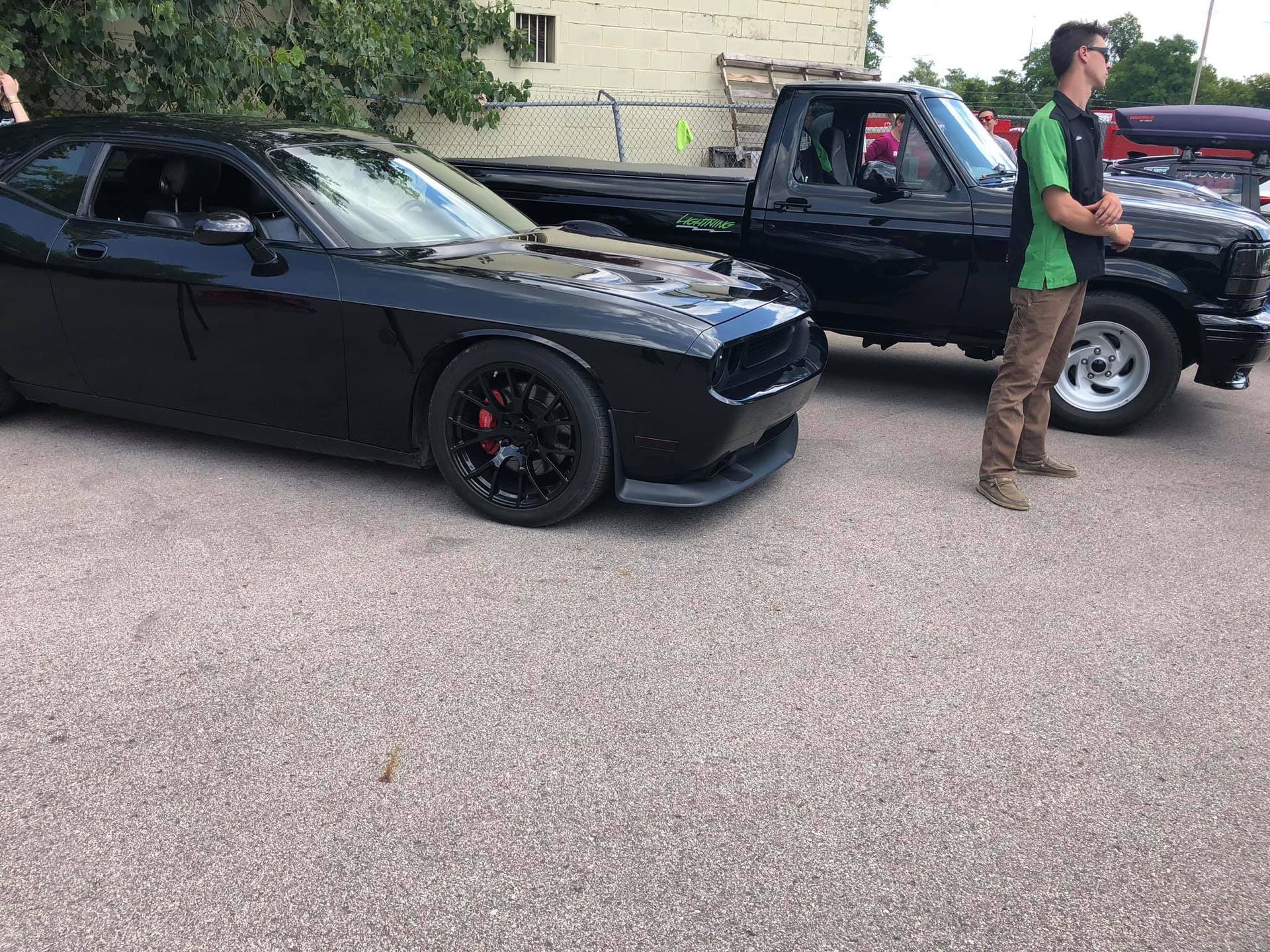 Black Dodge Challenger and truck parked at an outdoor car show, man standing nearby.