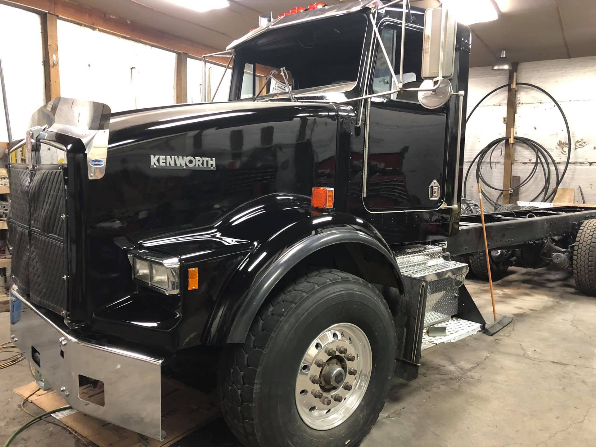 Black Kenworth semi-truck cab in a garage, with a chrome bumper and grill.
