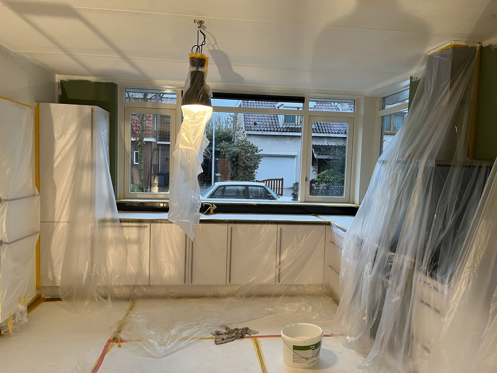 Schilderwerk keuken, kasten en ramen bedekt met plastic, met hangende werklamp.
