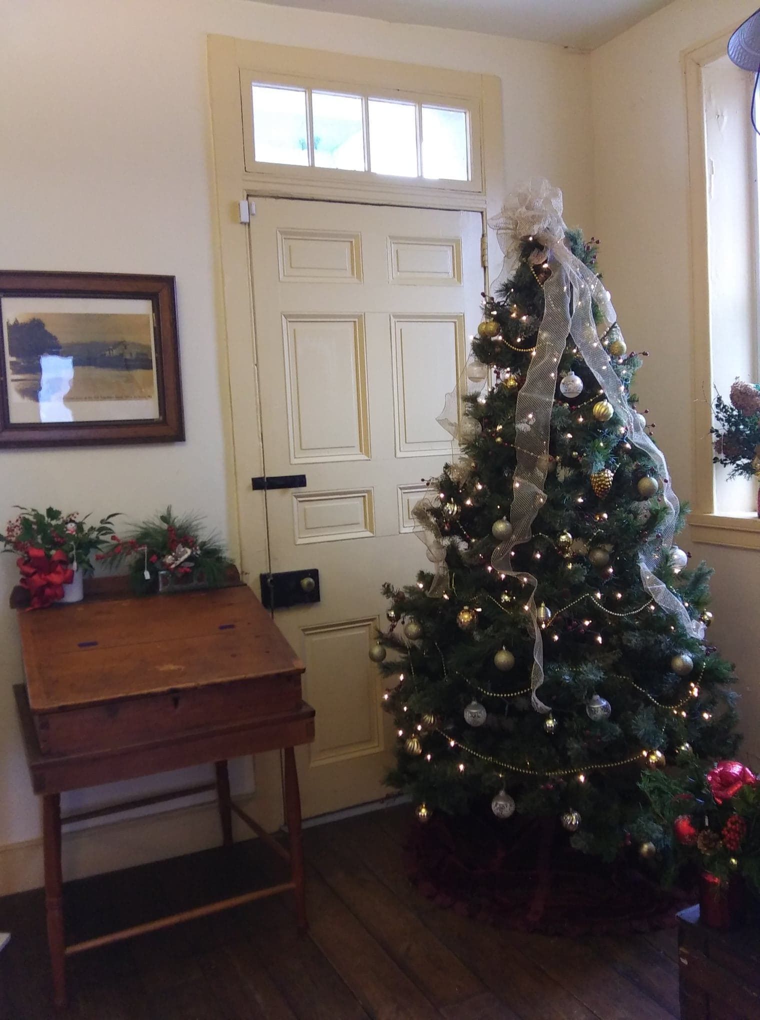 A Christmas tree decorated with gold and silver ornaments stands next to a door and a wooden desk in a room.